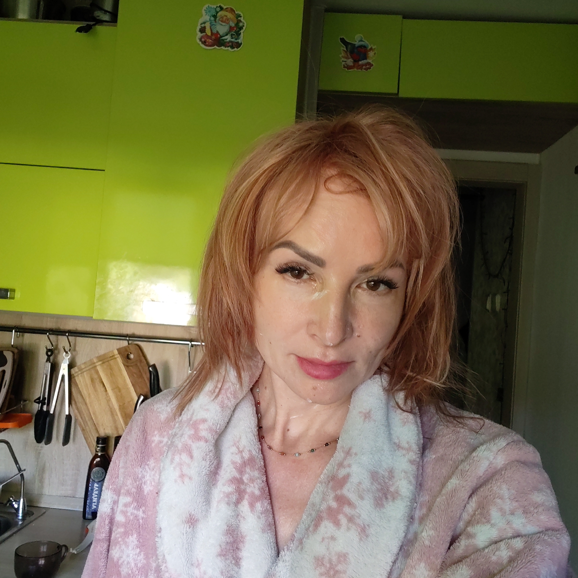 A woman is taking a selfie in a kitchen. She has reddish-blonde hair and is wearing a pink and white floral robe. There appears to be a facial mask on her face. The kitchen has bright green cabinets with stickers on the upper cabinets. Kitchen utensils and cutting boards hang on a rack. A bottle labeled "VLHVEYP" is visible. A mug sits on the counter next to a sink. A doorway is visible in the background. The location is Chaikovsky, Russia.