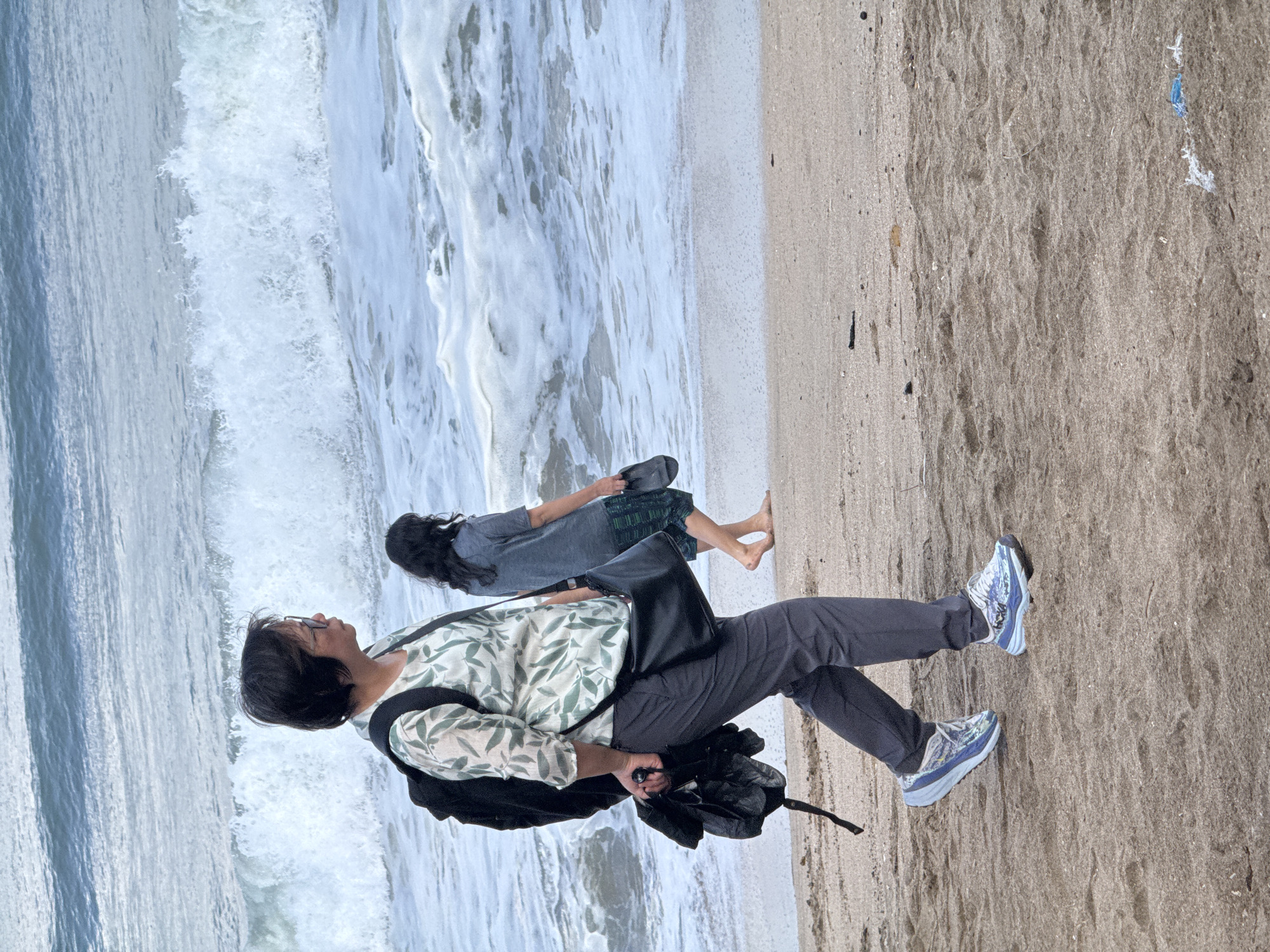 A woman with dark hair and glasses walks along a sandy beach in Kuta, Indonesia, with the ocean and waves behind her. She is wearing a white and green floral print shirt, dark pants, and light blue athletic shoes. A black backpack is slung over her left shoulder, and a black crossbody bag is worn across her body. She is holding a black umbrella in her right hand. Another person, partially visible, walks further down the beach in the background, wearing a grey t-shirt and shorts, carrying a dark sandal. The waves are breaking on the shore, and the sand is light brown with some scattered debris.