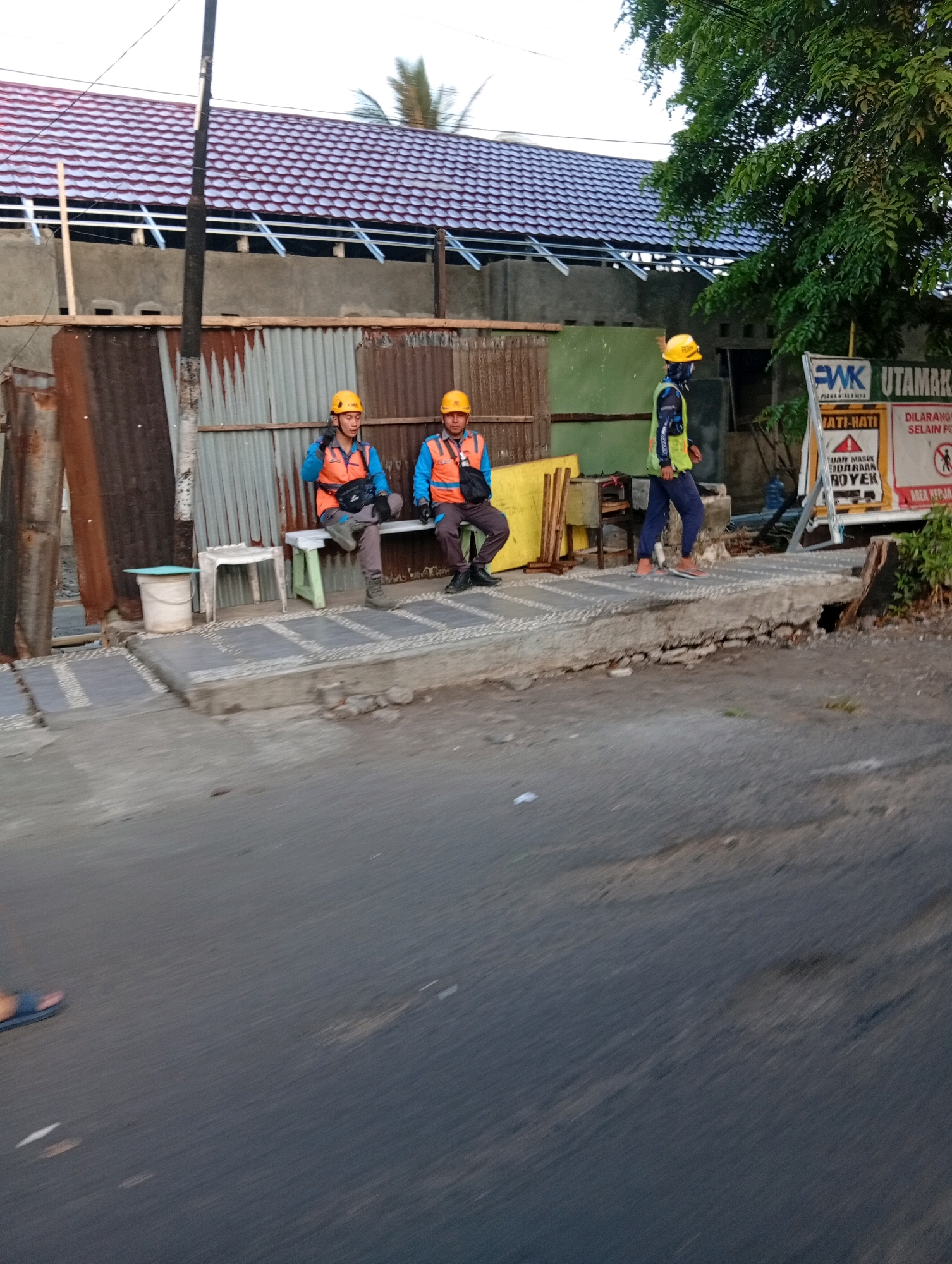 Three construction workers wearing yellow hard hats and orange safety vests are visible in the image. Two of them are sitting on a green and white bench, while the third is standing. The standing worker is wearing a blue shirt with a yellow and black safety vest, and dark blue pants. The two seated workers are wearing light blue shirts under their orange vests. One of them is holding a black bag. They are positioned on a concrete pathway next to a corrugated metal fence. Behind the fence, a building with a red tiled roof is under construction, with visible metal framing. A large tree with green foliage is on the right side of the image, partially obscuring a white sign with red lettering. The sign appears to be a warning sign with text in Indonesian. The scene appears to be an outdoor construction site in Manado, Indonesia, with a road in the foreground showing motion blur, suggesting the photo was taken from a moving vehicle.