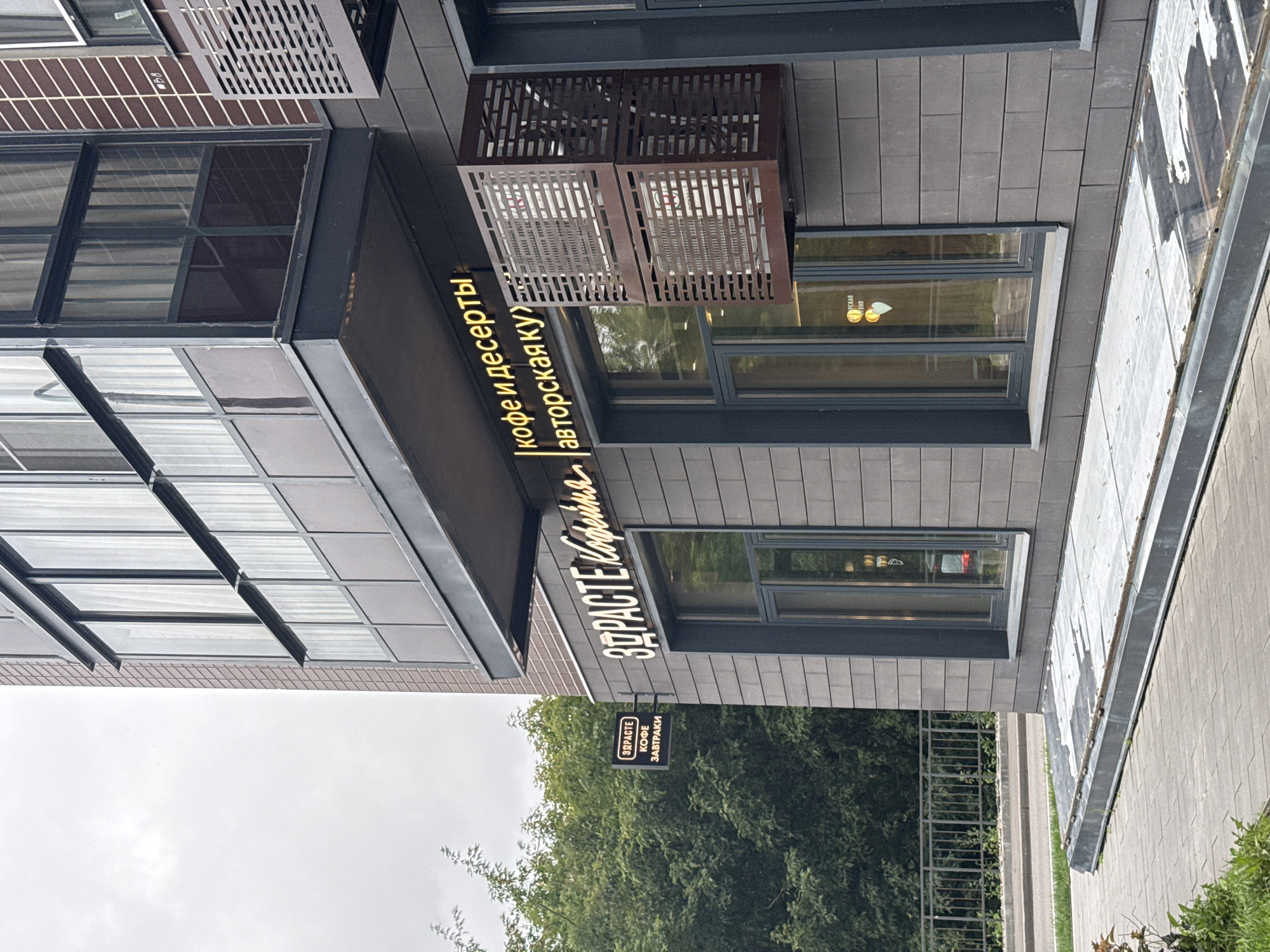 The image shows the exterior of a building with a sign that reads "ЗДРАСТЕ Кофейна". The sign is illuminated and has a cursive font. Below this sign, another sign reads "КОФЕ И ДЕСЕРТЫ авторская ку". There is also a smaller sign on a pole to the left with the text "ЗДРАСТЕ КОФЕ ЗАВТРАКИ". The building's facade is made of dark grey, brick-like tiles. There are large windows and what appears to be an air conditioning unit mounted on the wall, enclosed in a decorative metal grate. In the background, there is lush green foliage from trees. The ground in front of the building is paved with grey tiles. This scene is located in the city of Lyubertsy, Russia.