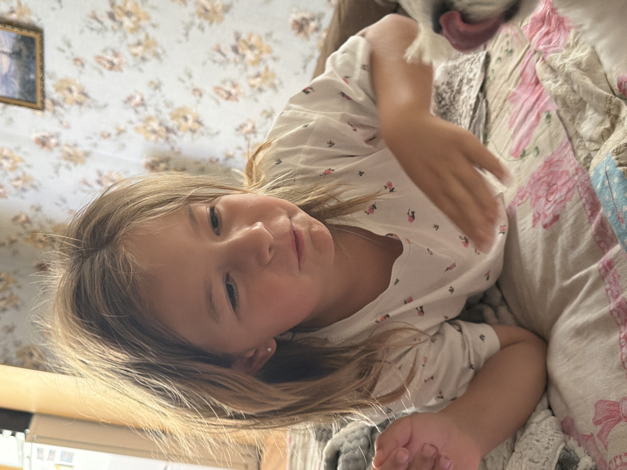 A young girl with blonde hair lies on a bed, her head tilted towards the camera. She wears a white t-shirt with a floral pattern. Her eyes are open, and she has a gentle expression. Beside her, a dog's snout and tongue are visible, suggesting the dog is interacting with her. The background shows floral wallpaper and a quilt with pink floral designs. The lighting appears to be natural, possibly from a window, suggesting daytime. The scene evokes a sense of comfort and affection between the girl and the dog. There are no visible text elements in the image.