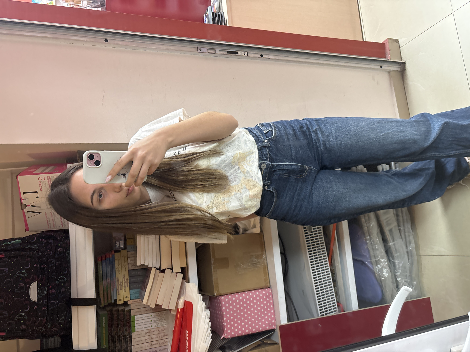 A woman is taking a selfie in a mirror. She is wearing a white t-shirt and blue jeans. She is holding a white smartphone with a pink case. She is standing in front of a shelf full of books, boxes, and other items. The shelf is located inside a store in Veliko Tarnovo, Bulgaria.