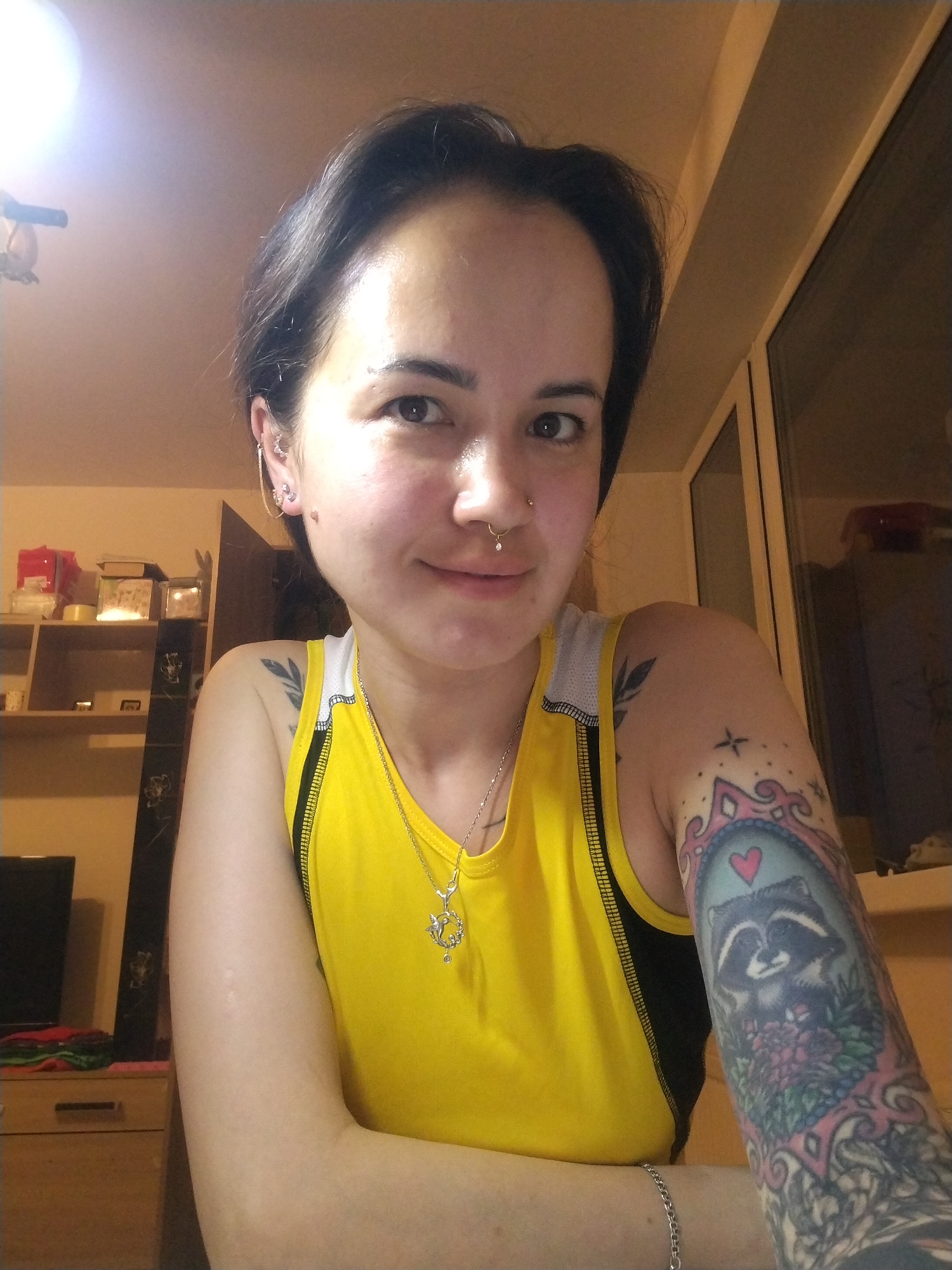 A woman with short dark hair and light skin is posing for a selfie. She is wearing a yellow tank top with black trim and a silver necklace. She has multiple piercings in her ears and a nose ring. Tattoos are visible on her left arm, one a raccoon in a frame with a heart above it, and on her left shoulder. She is standing indoors, in front of a window and a shelf with boxes. There are items on a dresser behind her. There is no evidence this image was taken in the city of Noviy, Russia.