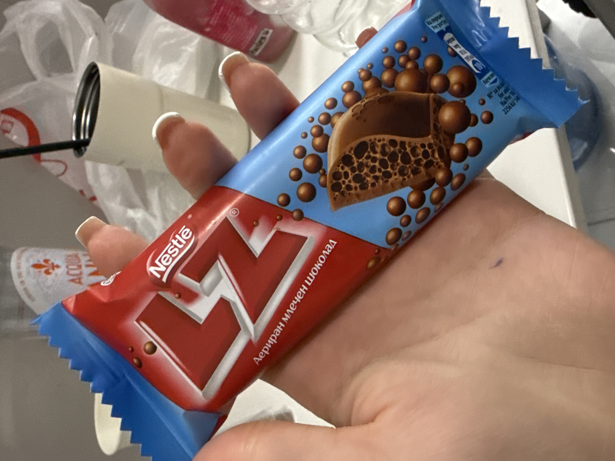 In this close-up, eye-level shot, a person's hand holds a Nestlé LZ chocolate bar. The chocolate bar has a red and blue wrapper adorned with the Nestlé logo, stylized "LZ" lettering, and images of chocolate bubbles and a cross-section of the aerated chocolate bar. The background is blurred, but hints of other objects, possibly in a domestic or retail setting, can be discerned, including a white cup, a thermal mug with a straw, and plastic packaging. The overall impression is one of a candid moment, with the focus firmly on the chocolate bar and the hand holding it. The lighting is bright, with a slight hint of artificiality.