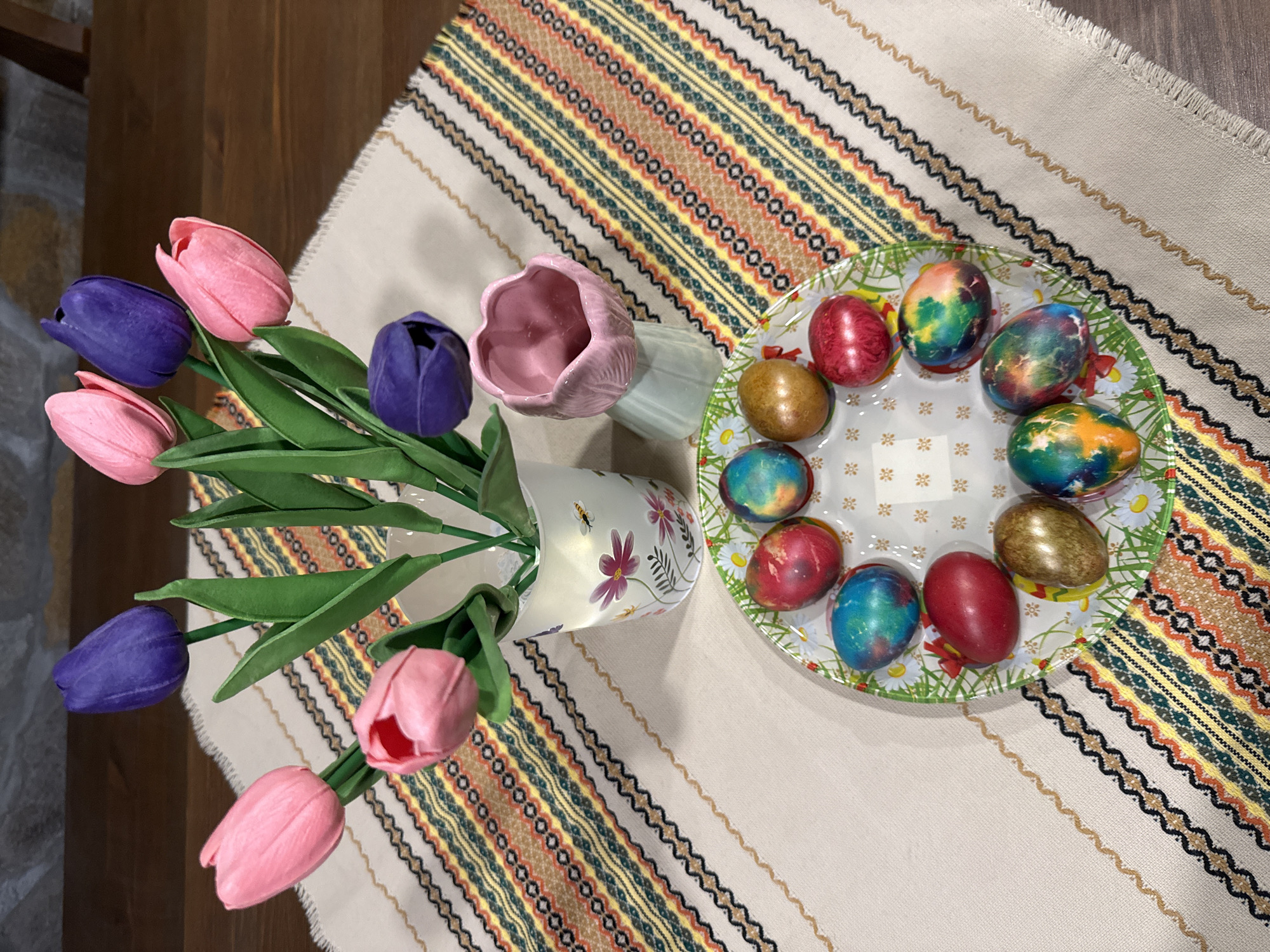 The image depicts a still life arrangement on a table, potentially indoors, within the city of Lukovit, Bulgaria. A patterned table runner, featuring geometric designs in beige, brown, yellow, green, and black, covers the surface. To the left, a vase filled with artificial tulips in pink and purple hues sits upright. Adjacent to the vase is a decorative pink ceramic piece, possibly another vase. A clear glass plate holding colored Easter eggs occupies the right side of the frame. The eggs exhibit a variety of colors, including red, gold, and multi-colored combinations of blue, green, and orange. The plate is decorated with a daisy pattern along its rim. The table is made of dark brown wood.