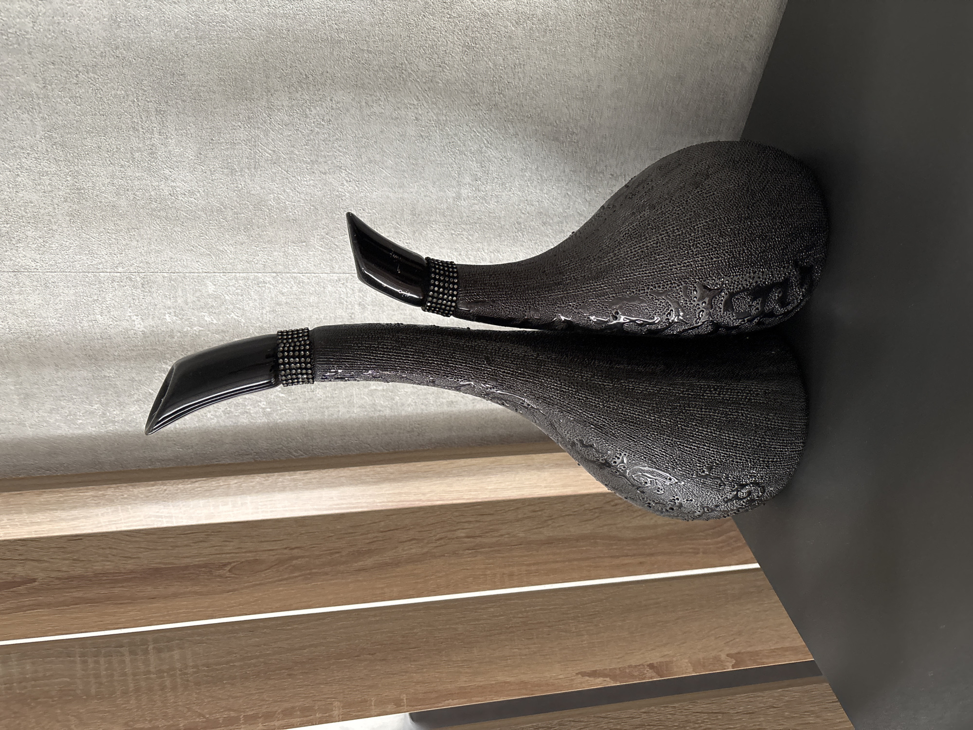 Two decorative vases, one taller than the other, are placed on a dark, flat surface. The vases are dark gray with a textured surface that appears to be covered in small raised dots. They have long, slender necks that curve upwards and outwards, culminating in a pointed black opening. Around the base of the neck of each vase, there is a band of small, sparkling beads. The larger vase is to the left and slightly behind the smaller vase. Behind the vases, there is a light gray, textured wall and to the left, a wooden partition with vertical planks. The lighting is even, with some subtle shadows cast by the vases. The scene appears to be an indoor setting, possibly a shelf or table in a home or display area, in a city not specified.