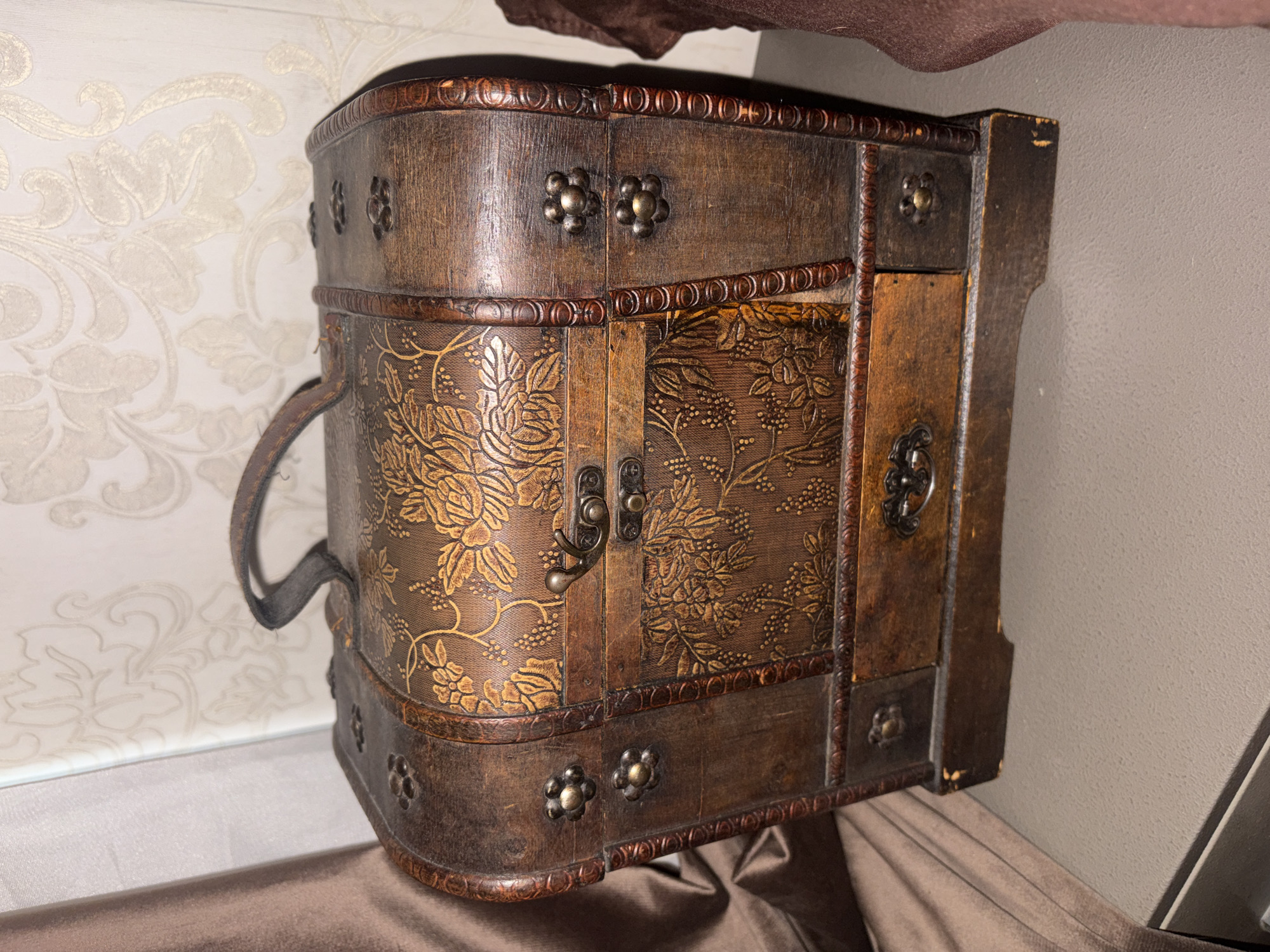 A close-up shot depicts a small, ornate wooden chest. The chest is brown with gold floral patterns and decorative metal studs. It features a hinged door with a latch and a carrying handle on the side. The surface beneath the chest is covered with a brown fabric. The background includes a patterned wallpaper and a plain wall. The image does not provide sufficient information to determine a location in Krasnoyarsk, Russia.