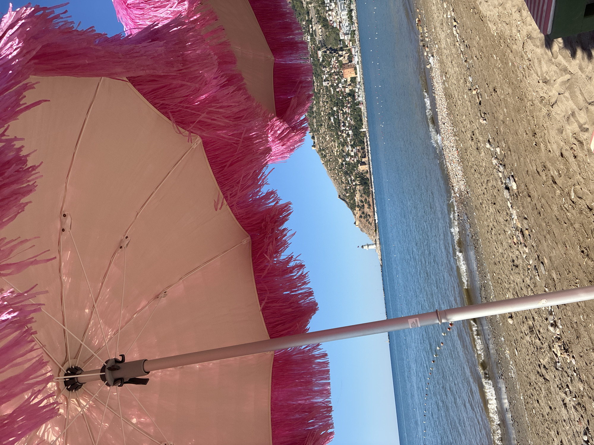 The image shows a beach scene in Alanya, Turkey, viewed from under a pink fringed beach umbrella. The umbrella's fabric is a light pink, and the fringe is a bright, vibrant pink. The pole of the umbrella is visible, extending down towards the sand. The beach is sandy with scattered pebbles. The sea is a calm blue, with small waves breaking on the shore. In the distance, across the water, a hilly coastline is visible, dotted with buildings and a prominent lighthouse on a hill. The sky is clear and blue. The overall impression is a sunny day at the beach.
