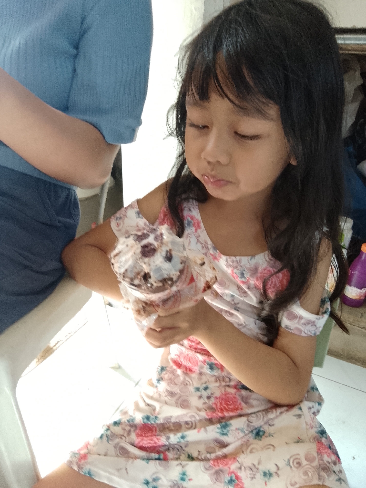 A young, dark-haired Asian girl is seated indoors, wearing a floral dress with cut-out shoulder sleeves. She is holding a plastic container filled with what appears to be chocolate ice cream or a similar dessert. Her eyes are lowered, and she seems to be in the process of eating.
To the left of the girl, a person wearing a blue shirt is partially visible. A white plastic chair is situated beside the girl.
Behind the girl, a purple bottle is visible on a shelf or surface.
The location is Davao City, Philippines.