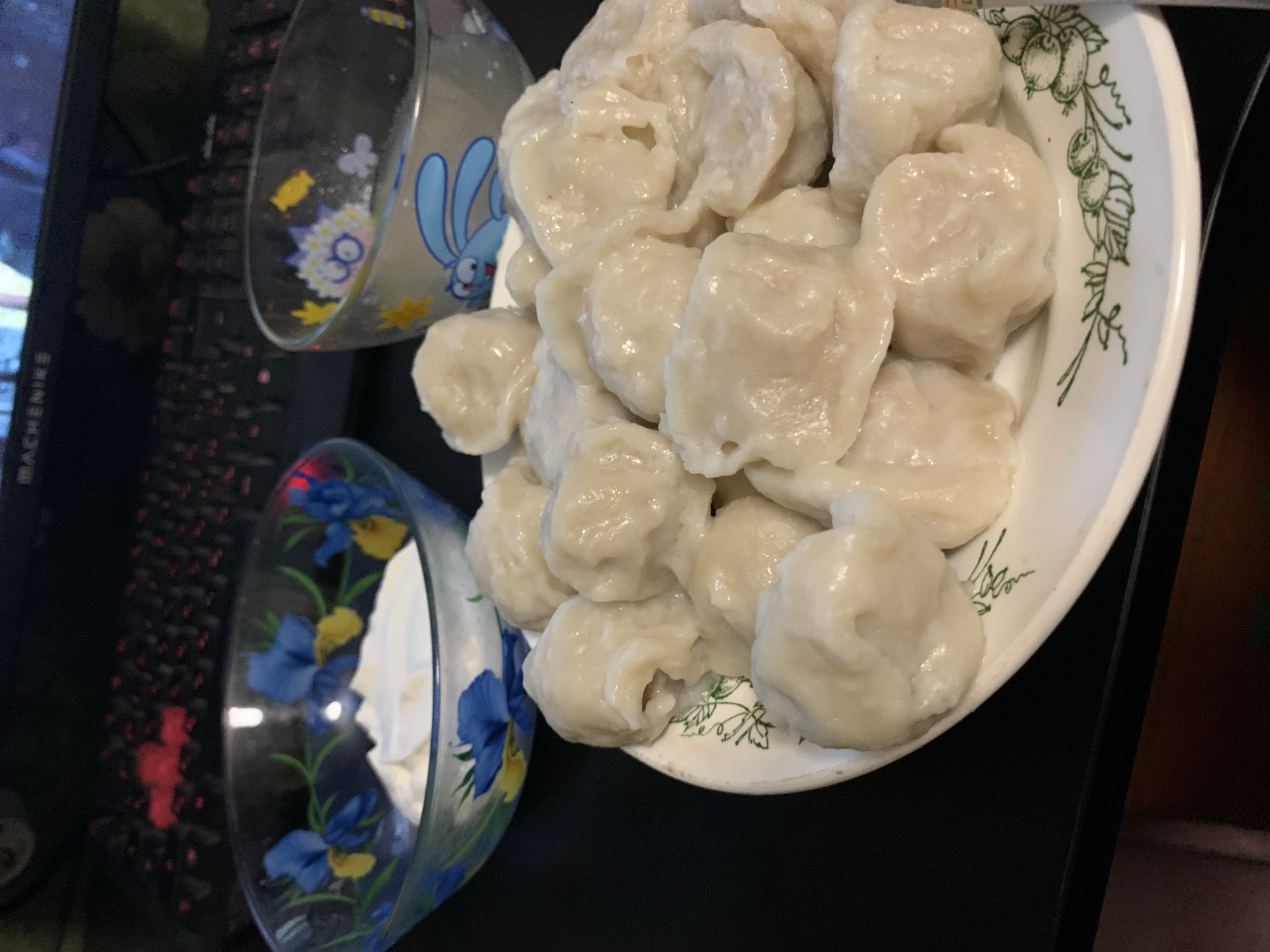 A plate of white dumplings sits on a black surface, with two decorative glass bowls behind it. The bowl on the left contains a white creamy substance, possibly sour cream or yogurt, and features a blue and yellow floral pattern. The bowl on the right is empty and decorated with a blue cartoon rabbit. A black keyboard is visible in the background. The overall scene appears to be indoors, possibly a dining area or kitchen. The dumplings are the primary subject, arranged in a pile on a white plate with a green floral rim.