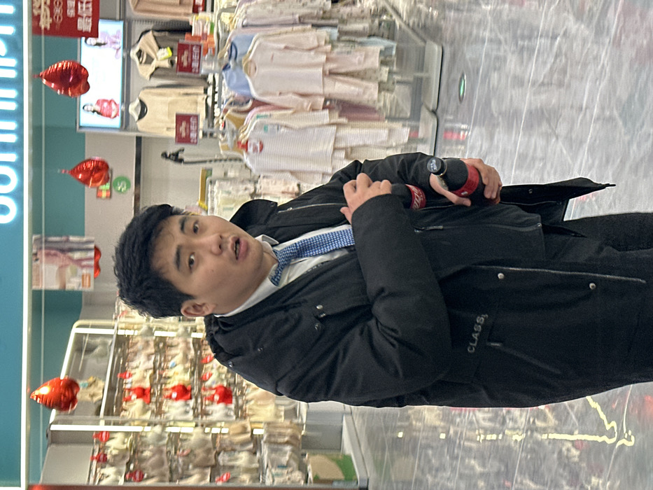 An adult male, appearing of East Asian descent, is captured in a brightly lit indoor retail environment within a department store section in Jiangjin, China. He stands slightly off-center, facing the camera with a slightly open-mouthed expression. He wears a black padded jacket with "CLASS," embroidered in white on the left side, over a white collared shirt and a disheveled blue patterned tie. In his right hand, he holds a dark bottle, identifiable as a Coca-Cola product by its red label and black cap. The floor is a reflective, polished light-colored tile. In the background, to the left, a display rack exhibits numerous beige brassieres. To the right, racks are filled with children's clothing, including light pink, white, and striped sleepwear. Red, teardrop-shaped foil balloons are suspended from the ceiling and interspersed among the displays. Overhead, a partially visible blue illuminated sign and promotional display boards featuring images of women and Chinese text are present.