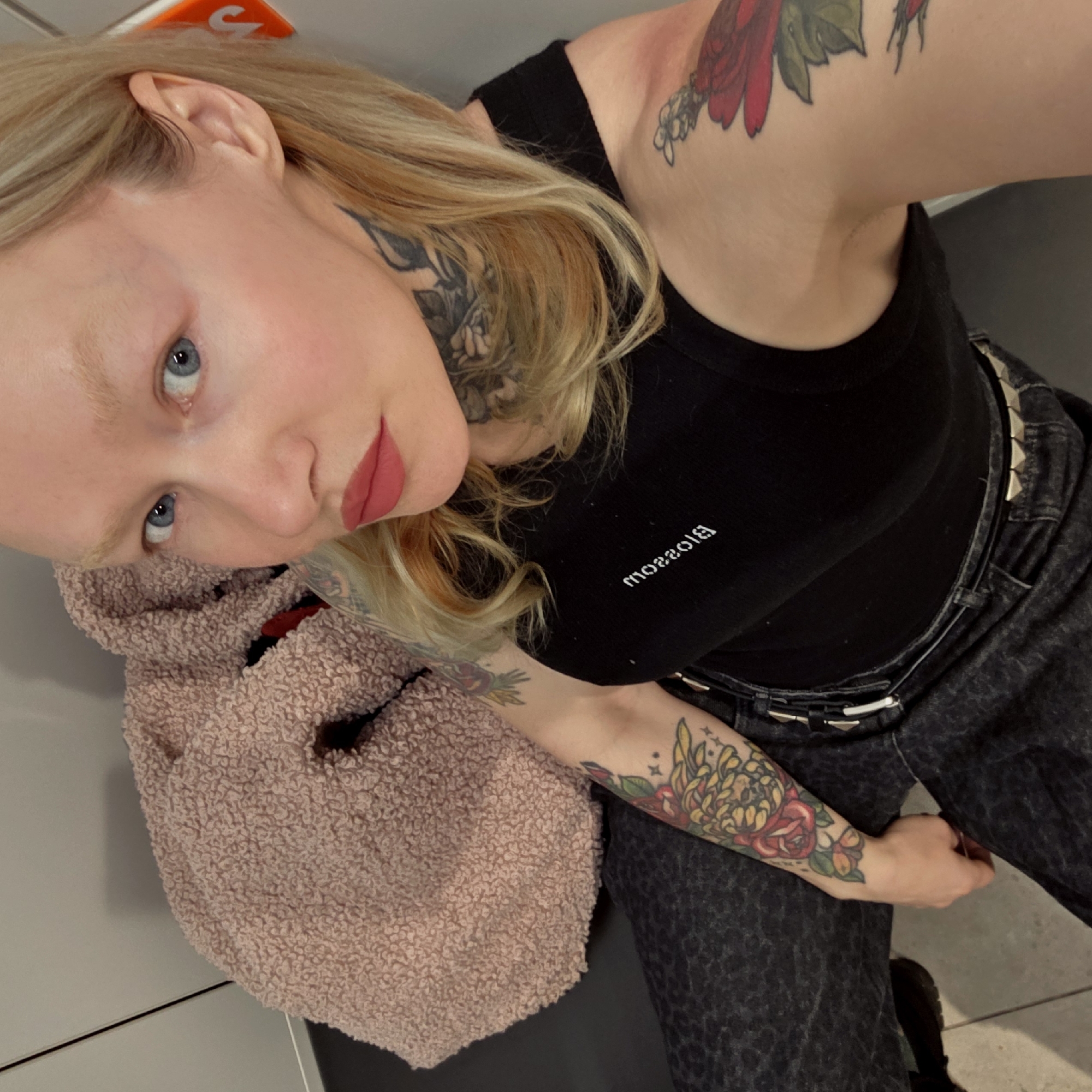 A person with light skin, blond hair, and blue eyes is looking up at the camera. They are wearing a black tank top with white text on it, and dark-colored pants with a silver belt. The person has multiple tattoos on their neck, arms, and shoulder. They are holding a fuzzy pink bag or item. The background appears to be a gray or white wall or surface. Without further information, the location in Saint Petersburg, Russia cannot be determined.