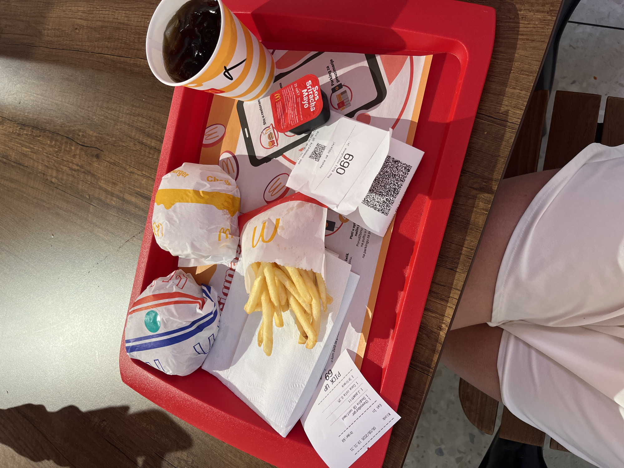 The image provides an overhead view of a McDonald's meal served on a red plastic tray, positioned on a wooden table. The meal consists of two burgers wrapped in paper (one with blue and red stripes, the other predominantly yellow), a carton of golden french fries, and a clear plastic cup filled with a dark carbonated beverage. A small red packet of Sriracha Mayo sauce is also present on the tray.
Multiple paper documents are visible. A primary receipt, dated "06/06/2023 19:31:31," details an order for "1 Cheeseburger," "1 Double Cheeseburger," "1 Coca Cola 0,25," and "1 Sr.fries," placed at a "Kiosk" for "Eat In." Another slip clearly displays "PICK UP 69," and a third receipt states "Vaš broj porudžbine je: 069" (Your order number is: 069) followed by "Hvala na poseti!" (Thanks for your visit!). The text on these documents and the tray liner, including phrases like "Šta u restoranu" (What in the restaurant) and "Parking za preuzimanje" (Parking for pickup), is in Serbian, confirming the location as a McDonald's restaurant in Belgrade, Serbia.
Beneath the table, the upper legs of an individual wearing light-colored shorts or a dress are partially visible, suggesting the person is seated at the table. The table itself is made of medium-brown wood. The floor visible in the background is a mix of wooden planks and lighter, possibly tiled, areas. A distinct shadow of a person, likely the photographer, is cast on the upper left section of the floor, indicating bright lighting conditions. A dark wooden chair is partially visible under the table. The scene captures a typical fast-food dining experience.