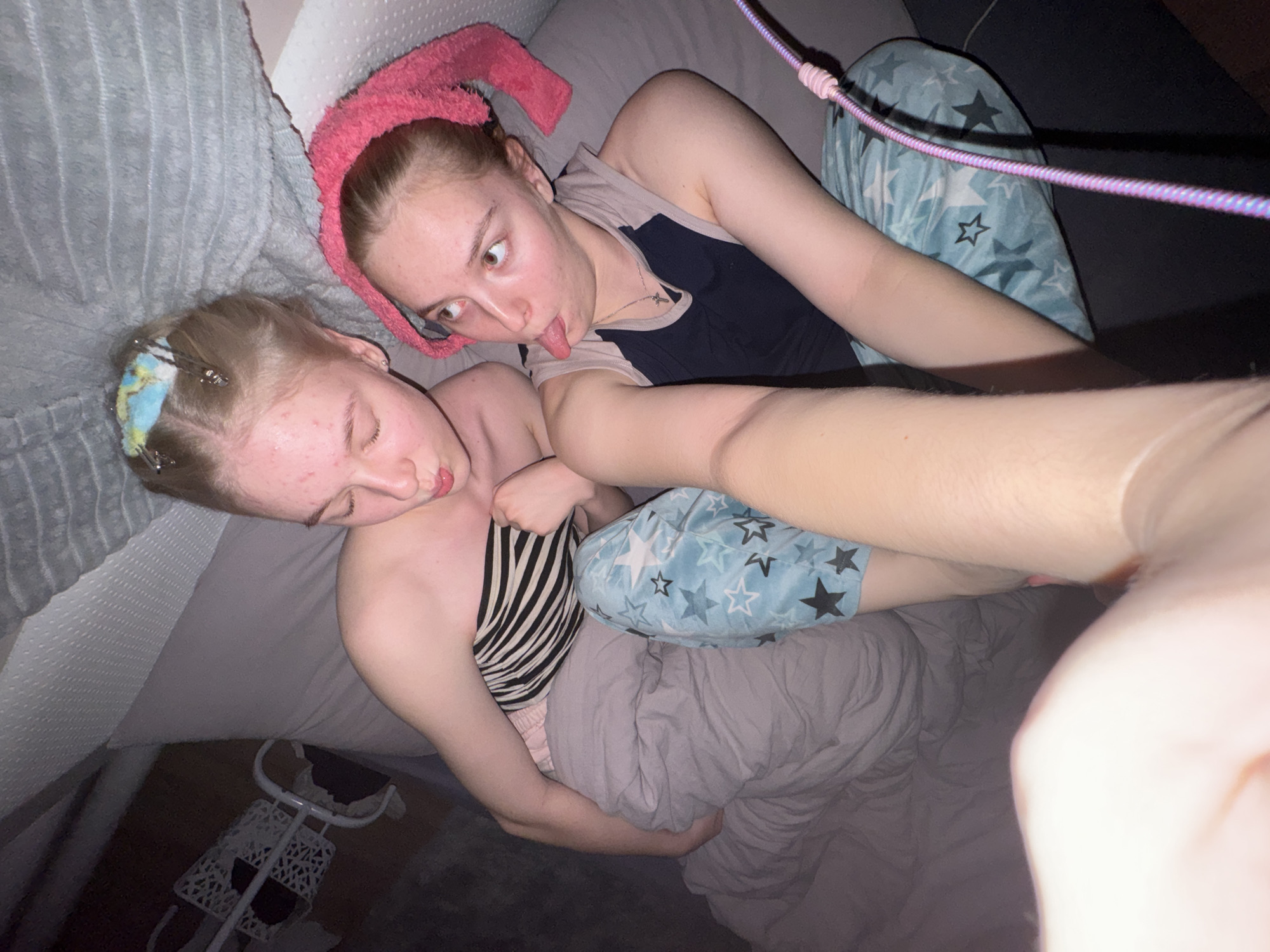 The media file shows two young females lying in a bed. The person in the foreground is leaning back, with their head on a pillow. They have blonde hair with a blue and yellow headband and hair clips. They are wearing a black and white striped top and pink shorts. The other person is lying on their back, with their head on a pillow covered by a pink towel. They have blonde hair and are wearing a dark blue tank top and light blue pajama pants with white and black stars. Their tongue is sticking out, and they are looking at the camera. The setting appears to be indoors, possibly a bedroom, with blankets and pillows visible. A white cart with a woven design is partially visible in the lower left corner of the image. The lighting suggests it is nighttime or a dimly lit room. The overall mood is playful and casual. There is no visible text in the image.