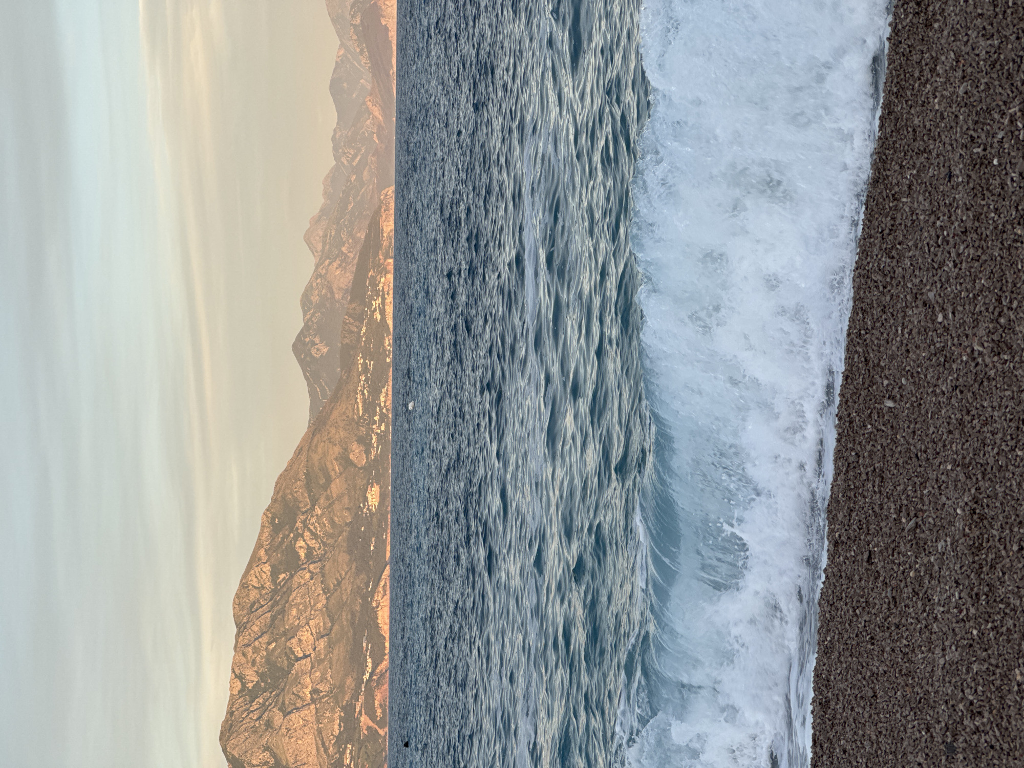 The image depicts a seaside landscape at Prijevor, Montenegro. In the foreground, a pebble beach is visible with small, dark-colored stones. A wave is breaking onto the shore, characterized by white foam and translucent blue water. The sea itself is a deep blue, with small ripples and crests on its surface. In the midground, a thin line of land is visible, featuring rocky hillsides dotted with small structures, suggesting a coastal settlement. The background is dominated by a range of majestic mountains, their slopes illuminated by the warm, soft light of what appears to be either sunrise or sunset. The sky above is a pale, hazy blend of light blue and pastel orange, with subtle cloud formations. There are no discernible people in the immediate foreground or midground. Two dark shapes are faintly visible in the water, one on the far left and another further to the right, possibly indicating distant figures or objects. A small white object, possibly a bird or a buoy, is also present in the water on the right side. The overall scene conveys a sense of natural beauty and tranquility.