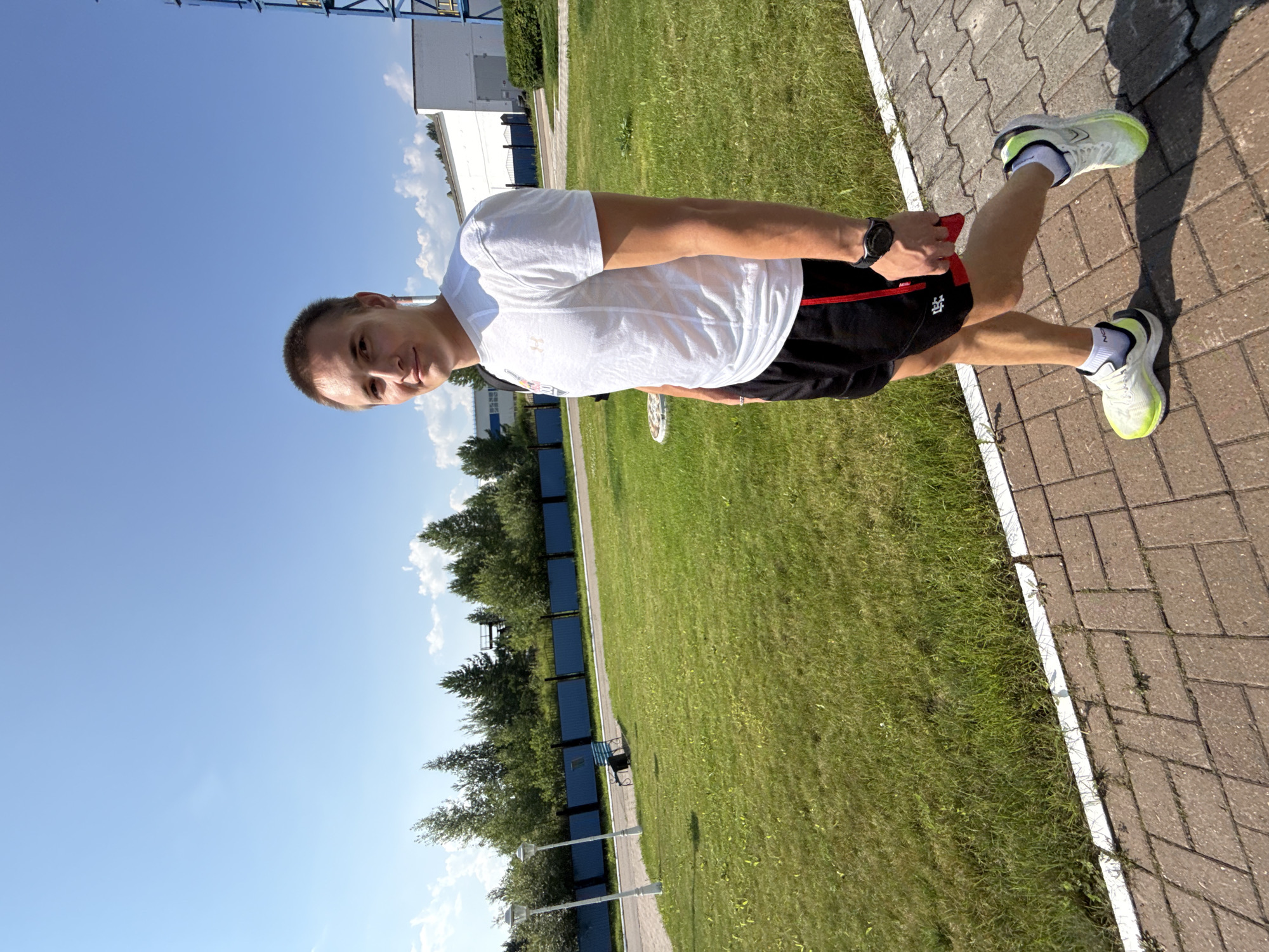 A young man with short brown hair stands on a paved walkway with a grassy area to his left. He is wearing a white t-shirt with a logo on the left chest, black athletic shorts with red trim, white socks, and neon yellow athletic shoes. He is looking at the camera with a slight smile. In the background, there is a blue fence, trees, and a white building with a tall blue structure next to it. The sky is clear and blue with a few white clouds. The image appears to have been taken outdoors in the city of Chaikovsky, Russia.