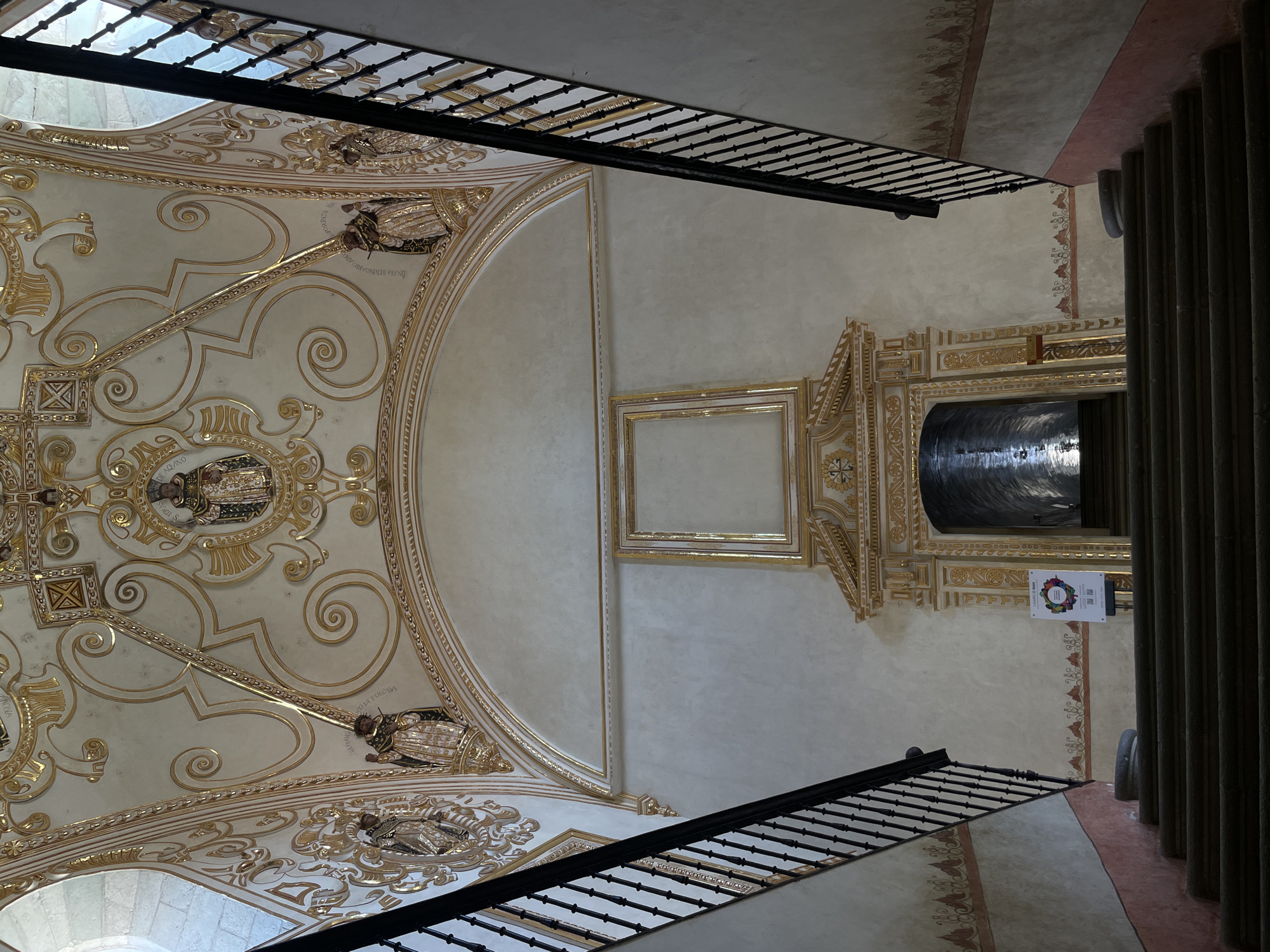 The image depicts the interior of a building in Oaxaca City, Mexico. The view is angled upwards, showcasing an ornate ceiling with elaborate gold detailing and patterns. The ceiling features a curved section adorned with decorative swirls and portraits. A plain square frame is visible, adjacent to an elaborate structure with a dark, reflective cylindrical element. A small sign with colorful graphics hangs nearby. Two sets of black metal railings extend diagonally across the image, framing the scene. A row of vertical pillars is visible on the right edge of the frame. The overall impression is of a historic and richly decorated space.