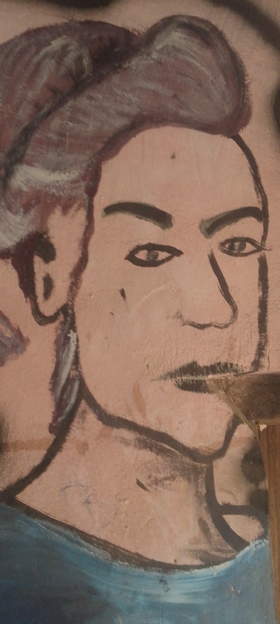 A close-up shot of a mural painted on a wall, depicting a face. The face is rendered with thick, dark outlines and minimal shading, giving it a graphic, almost primitive quality. The subject's hair is a dark, reddish-brown with highlights of white and gray, styled in a swept-back manner. The eyes are depicted with simple dark circles for pupils and dark lines for the eyelids and eyebrows. The nose is sharply defined, and the mouth is a thin, dark line with subtle shading. A mole is visible on the left cheek. The skin tone appears to be a light peachy-brown. The mural extends beyond the face, with hints of another figure to the left and a blue garment covering the lower portion of the depicted subject. The wall itself is a light, textured brown. A wooden object, possibly part of a structure or frame, juts into the lower right corner of the frame. The artwork is located on a wall in Jalingo, Nigeria.