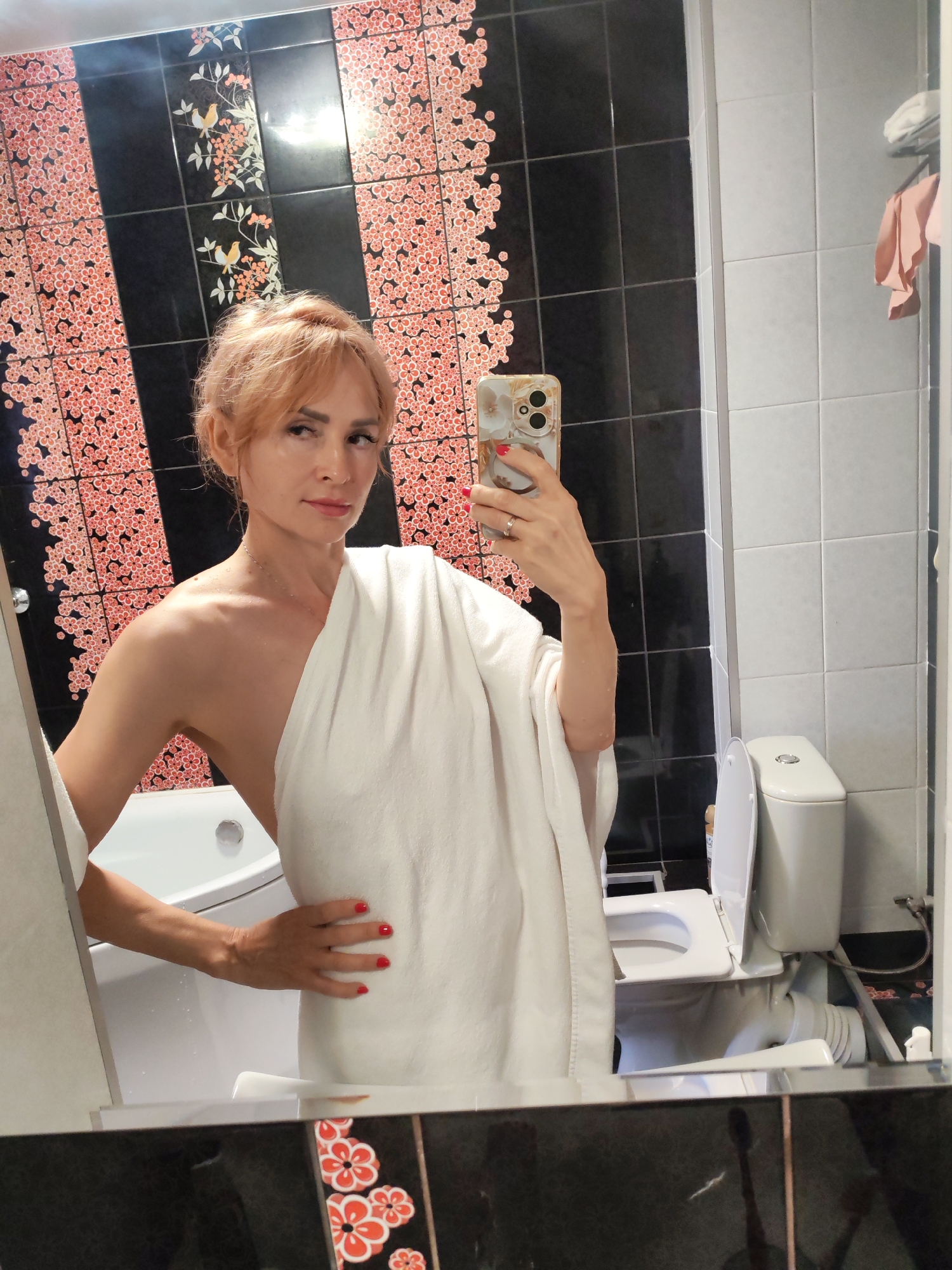 A woman is taking a mirror selfie in a bathroom. She is wearing a white towel draped over her body. Her hair is styled up and she has makeup on. In the reflection, we can see a white bathtub, a toilet with the lid up, and a tiled wall with a floral pattern. The bathroom appears to be well-lit. The woman's expression is neutral. There is no visible text in the image.