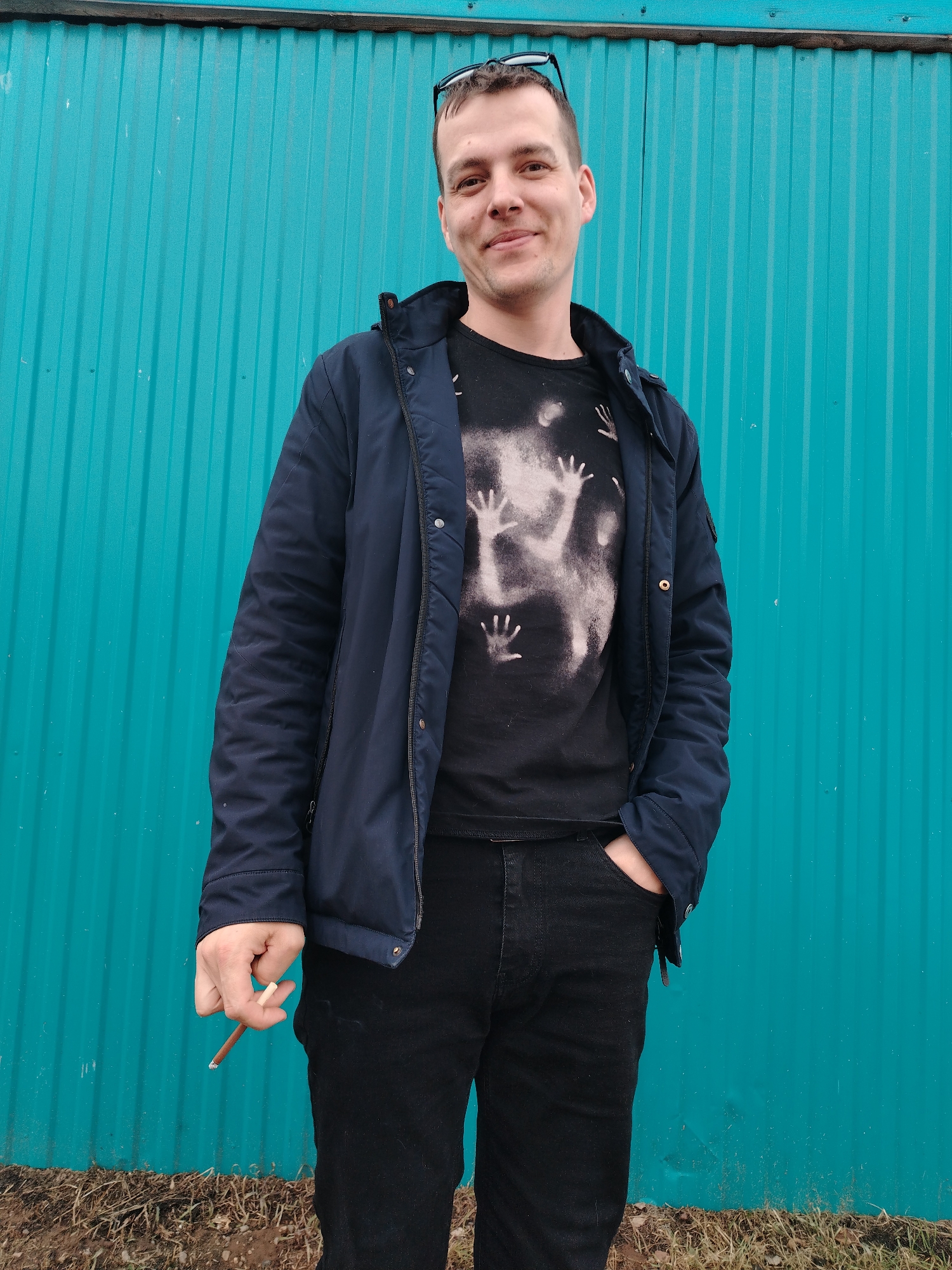An adult male stands outdoors in Nizhnyaya Gar', Russia, positioned against a vibrant teal corrugated metal wall. The subject is light-skinned, with short brown hair, and exhibits a faint smile directed towards the viewer. Sunglasses are perched on top of his head. He wears an unzipped dark blue jacket over a black t-shirt, which features a prominent bleached or light-colored graphic design depicting multiple upward-reaching hand outlines. He also wears dark-colored trousers, with his left hand inserted into a pocket. His right hand hangs loosely, holding a lit cigarette or blunt, from which a slight wisp of smoke or ash is visible near the tip. The ground directly in front of the corrugated wall consists of brown soil interspersed with sparse dry grass and twigs. A horizontal wooden beam is visible along the top edge of the corrugated wall.