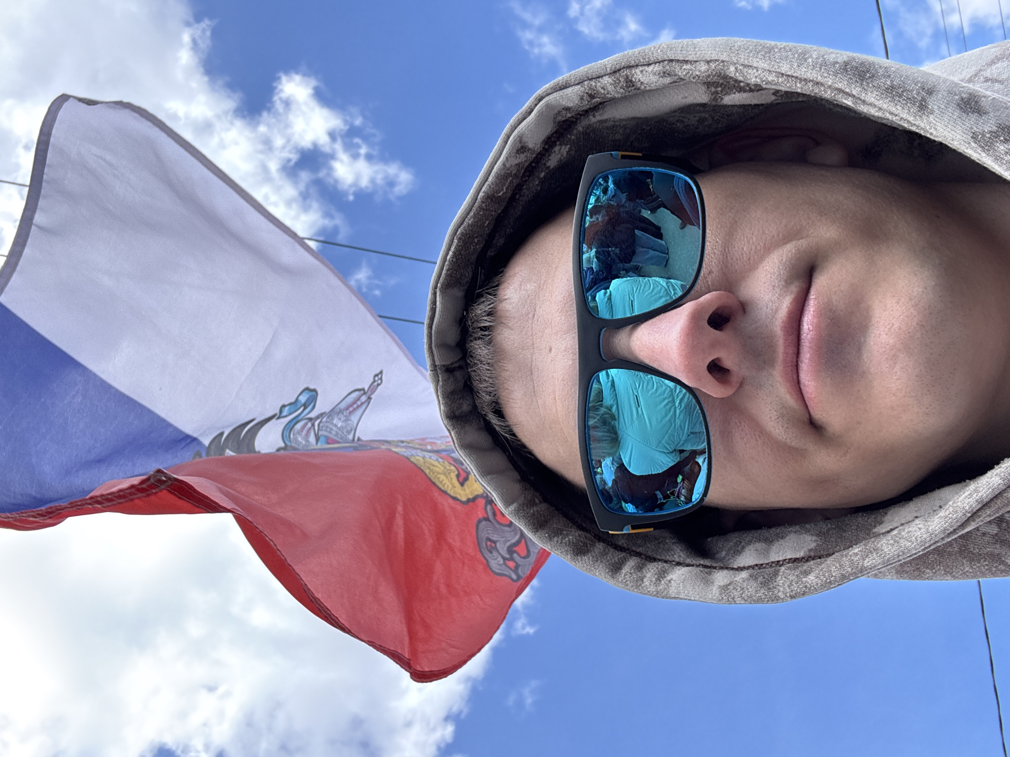A man with short hair, wearing a grey hooded sweatshirt and blue mirrored sunglasses, is shown in a close-up, low-angle view. The sky is visible in the background, partly covered with white clouds. A Russian flag is prominently displayed to the left of the man, with its white, blue, and red bands visible. The flag features an emblem on the blue stripe. The reflection in the sunglasses appears to show an interior space, possibly with clothing or fabric. The image was taken in Chuprino, Russia.