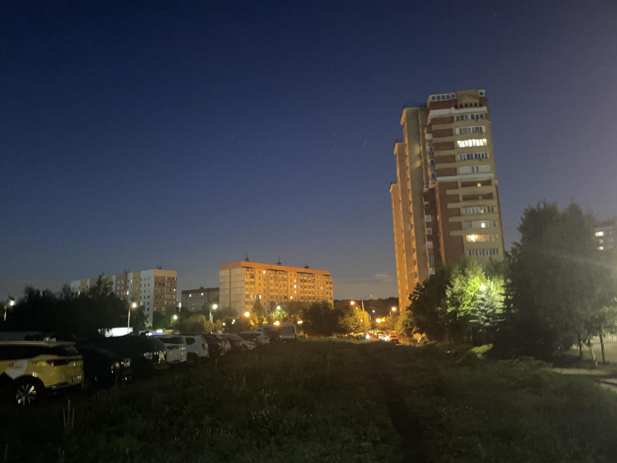 The image depicts a nighttime scene in a residential area of Krasnogorsk, Russia. In the foreground, a patch of unkempt grass and vegetation leads to a parking lot filled with various cars. A yellow car with white markings is visible on the left side of the parking lot. Behind the parking lot, several multi-story apartment buildings are illuminated from within, suggesting that it is evening or night. The largest building on the right is a tall, modern apartment complex with a reddish-brown facade and numerous windows, some of which are lit. To its left, a longer, lighter-colored apartment building is also visible, with lit windows. Trees and streetlights are scattered throughout the scene. The sky is a deep, dark blue, with a few faint streaks that might be atmospheric phenomena or distant clouds. The overall atmosphere is one of a typical urban residential environment at night.