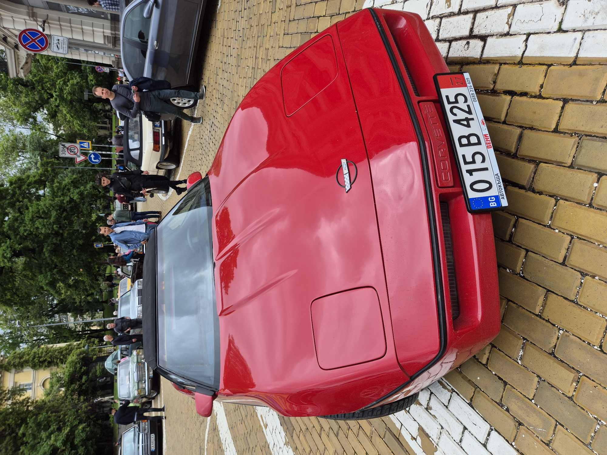 The image shows a red Corvette parked on a brick pedestrian area in Sofia, Bulgaria. The car faces the camera and is angled slightly to the right. The car's license plate is prominently displayed. People and other cars are visible in the background, suggesting the car is part of a gathering of classic cars. Street signs are also visible in the background, confirming the location is in Sofia.