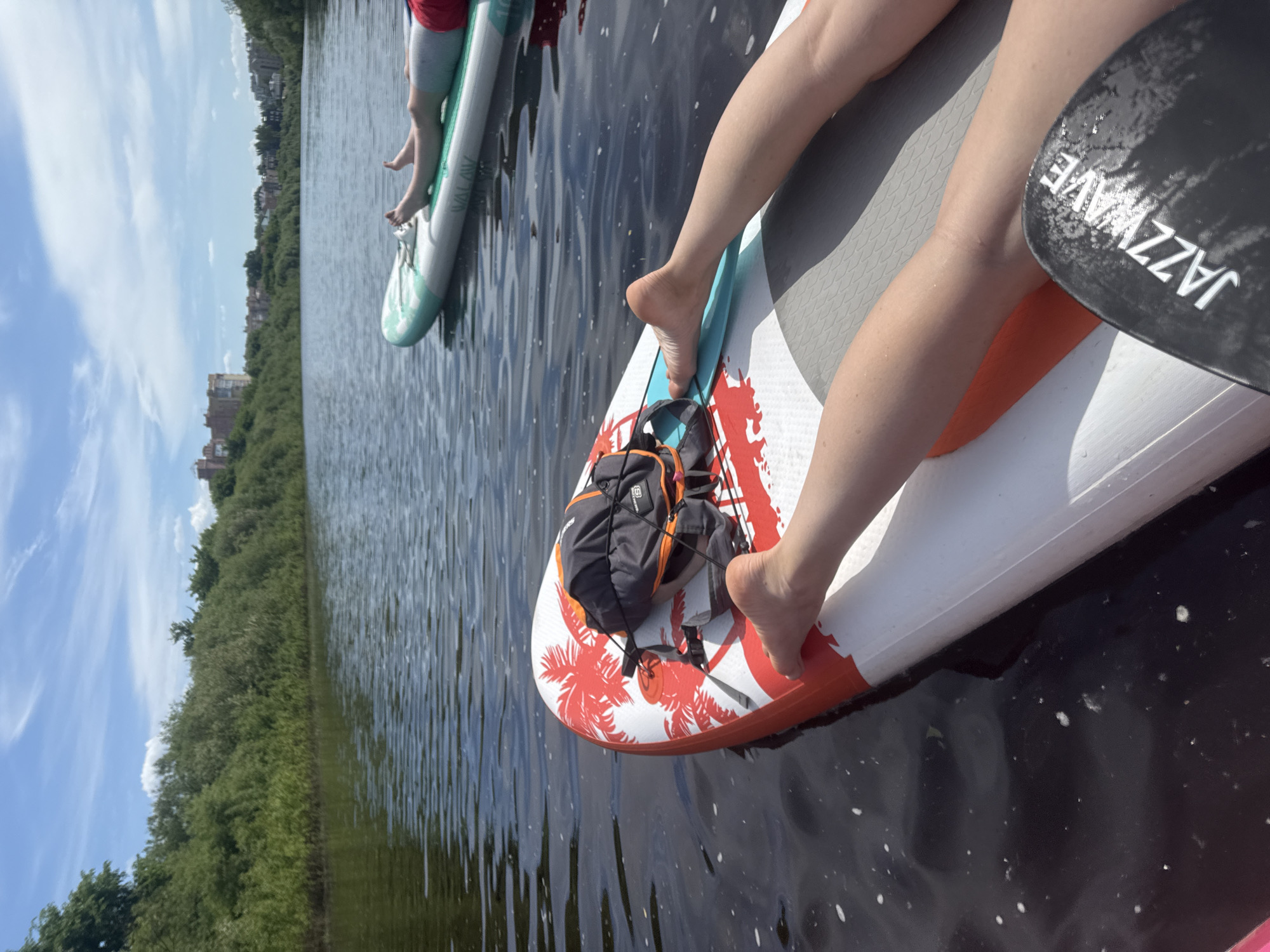 Two individuals are paddleboarding on a river under a bright blue sky with scattered white clouds. In the foreground, a person's bare legs and feet are extended on a white paddleboard featuring red palm tree graphics. A black and orange backpack is secured with bungee cords near the front of this board, and a black paddle blade with "JAZZWAVE" branding is partially visible in the lower right. Further into the frame, a second person's lower legs, clad in light-colored shorts and a red top, are visible as they recline on a light green and white "WALAW" branded paddleboard. The dark river water shows minor ripples and some small white foam specks. Both banks of the river are densely covered with green trees and bushes. In the distant background, beyond the treeline, several multi-story buildings of urban design are visible on the far riverbank. The setting is consistent with the Vologda River within the city of Vologda, Russia, characterized by its mix of natural riverside vegetation and adjacent urban development.