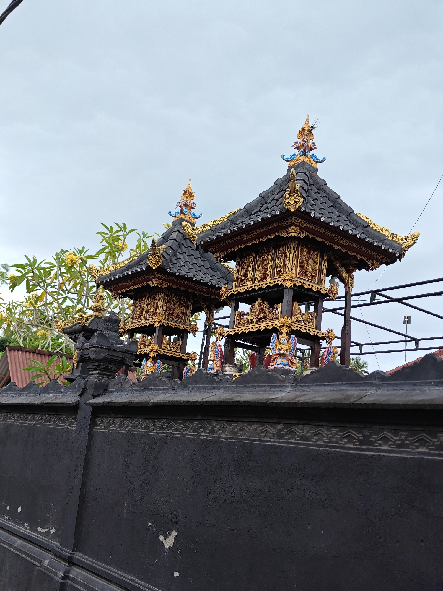The image shows two small, ornate pavilions with tiered roofs and intricate gold detailing, likely Balinese shrines, situated behind a dark, decorative wall. The location is in Nusa Penida, Indonesia. The pavilions feature dark-tiled roofs, gold ornamentation, and small, colorful sculptures visible at their base. A black wall with a decorative band runs across the foreground, partially obscuring the view of the structures. The sky is overcast. A tree with yellow flowers is visible to the left, and what appears to be a simple red roof is seen in the background. Metal construction support is behind the pavilions.