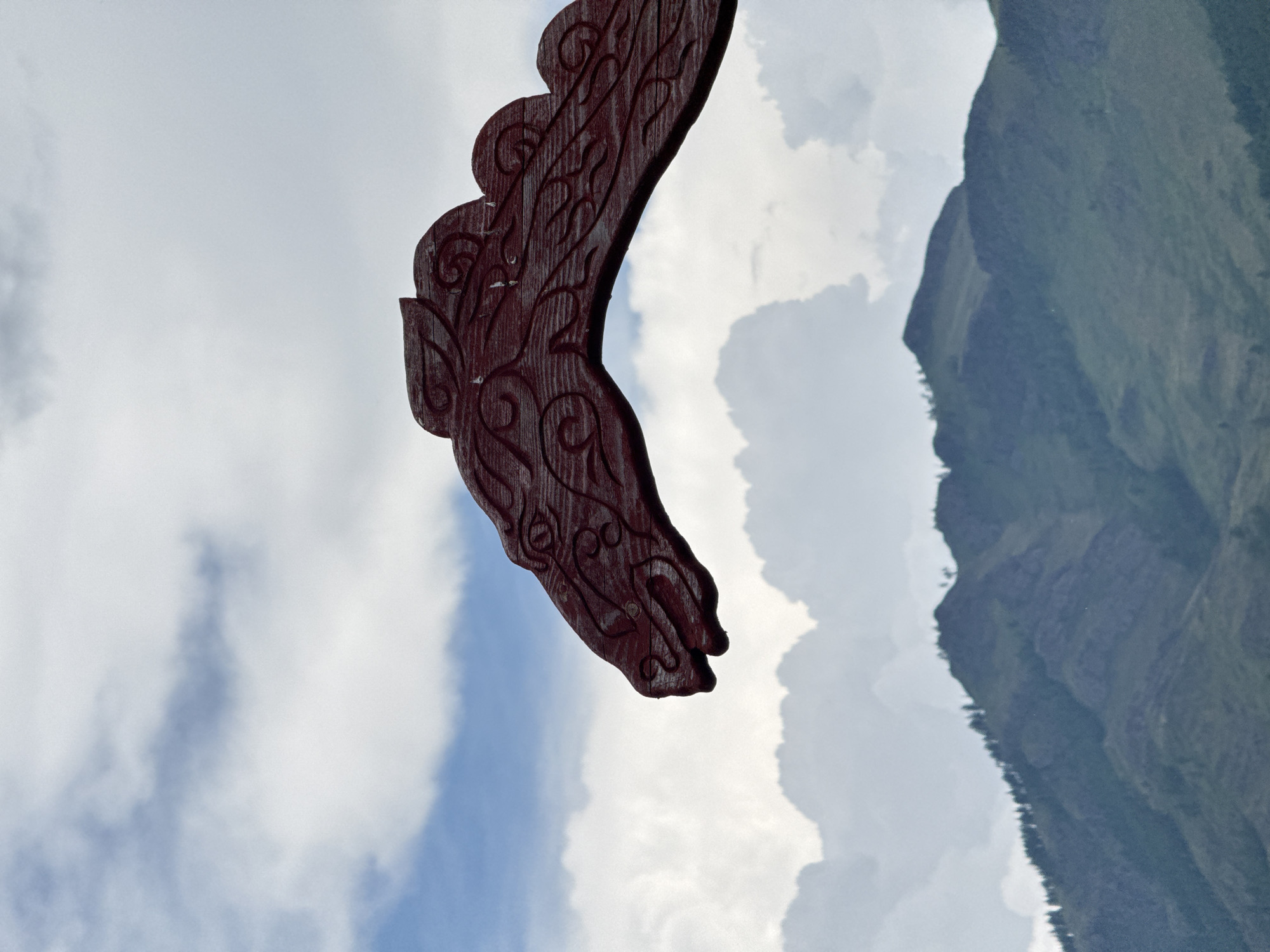 An outdoor, eye-level shot captures a detailed, carved wooden dragon head, painted a deep red, extending from the right side of the frame, its snout pointing left. The dragon's head features intricate swirling patterns carved into its surface, and its eye is a circular indentation. The background is a layered landscape of sky and mountains. The upper portion of the sky is a pale blue with wispy white clouds. Below the sky, a range of rugged, green mountains stretches across the horizon, dotted with dark green trees on their slopes. The overall lighting suggests it is daytime, with diffused light illuminating the scene. The image is sharp and clear, focusing on the dragon head's texture and the mountain's form. The location is Anchyl-Chon, Russia.