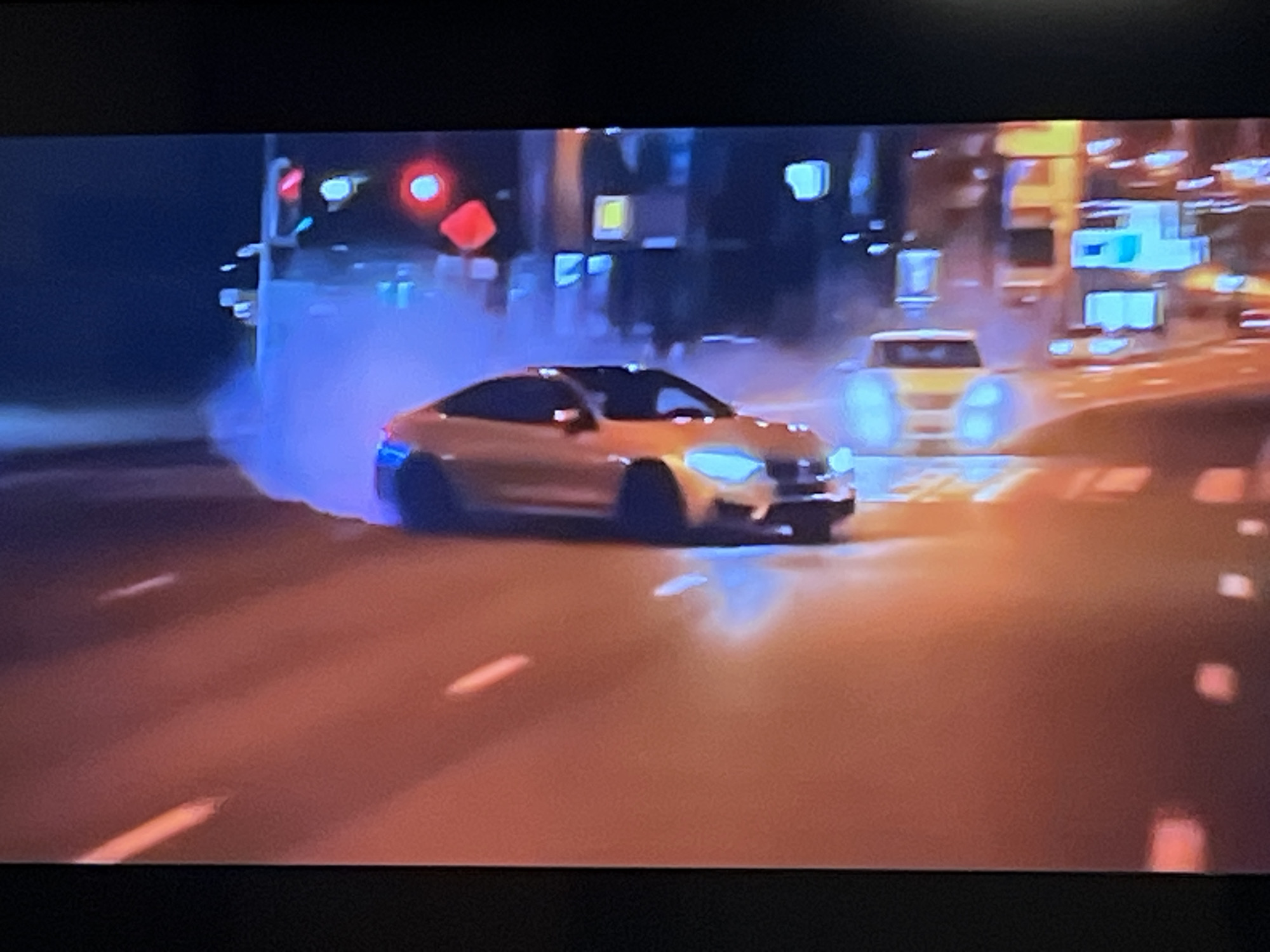 This image depicts a nighttime street scene with a prominent golden-colored sports car drifting, creating a cloud of blue smoke around its rear tires. The car is in motion, leaning into a turn on a wet or slick road surface that reflects the surrounding lights. In the background, there are out-of-focus city lights, including traffic signals that appear to be red and possibly other illuminated signs or building lights. A second, lighter-colored vehicle is visible further down the road. The overall atmosphere suggests a high-speed, dynamic event. The image does not contain any discernible elements that can be used to identify the specific location as Kharkiv, Ukraine.