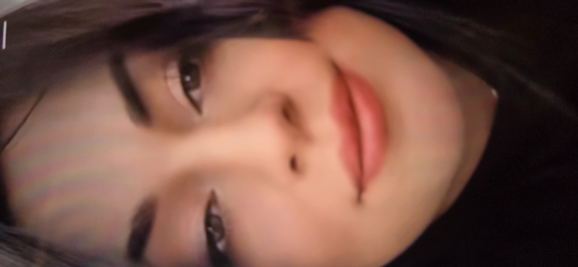 The image is a close-up, low-resolution, pixelated photograph of a young woman's face, focusing on her eyes and mouth. She has dark hair and appears to be wearing light-colored makeup on her eyes and pink lipstick. There is a slight overexposure on her forehead, which has a rainbow-like color cast. The background is dark and out of focus, obscuring any details about the environment. It is impossible to determine the location within the city of Kurgan, Russia, from this image.