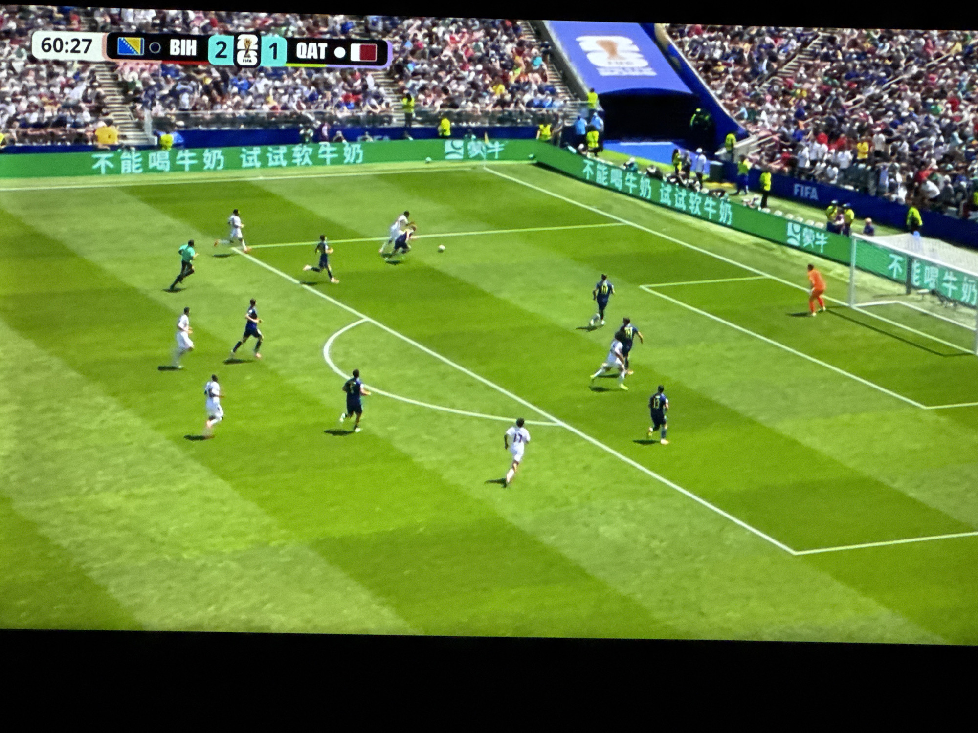 The image captures a soccer match during the day, with players in white and dark blue uniforms on a green field. The scoreboard at the top left indicates the time as 60:27, with "BIH" leading "QAT" 2-1. Advertising banners with Chinese text are visible along the sidelines. A stadium with a large crowd is in the background. The architecture and stadium design are not distinct enough to pinpoint a specific city or country without further information, but the mention of "Russia" in the prompt suggests it might be a venue from the 2018 FIFA World Cup.