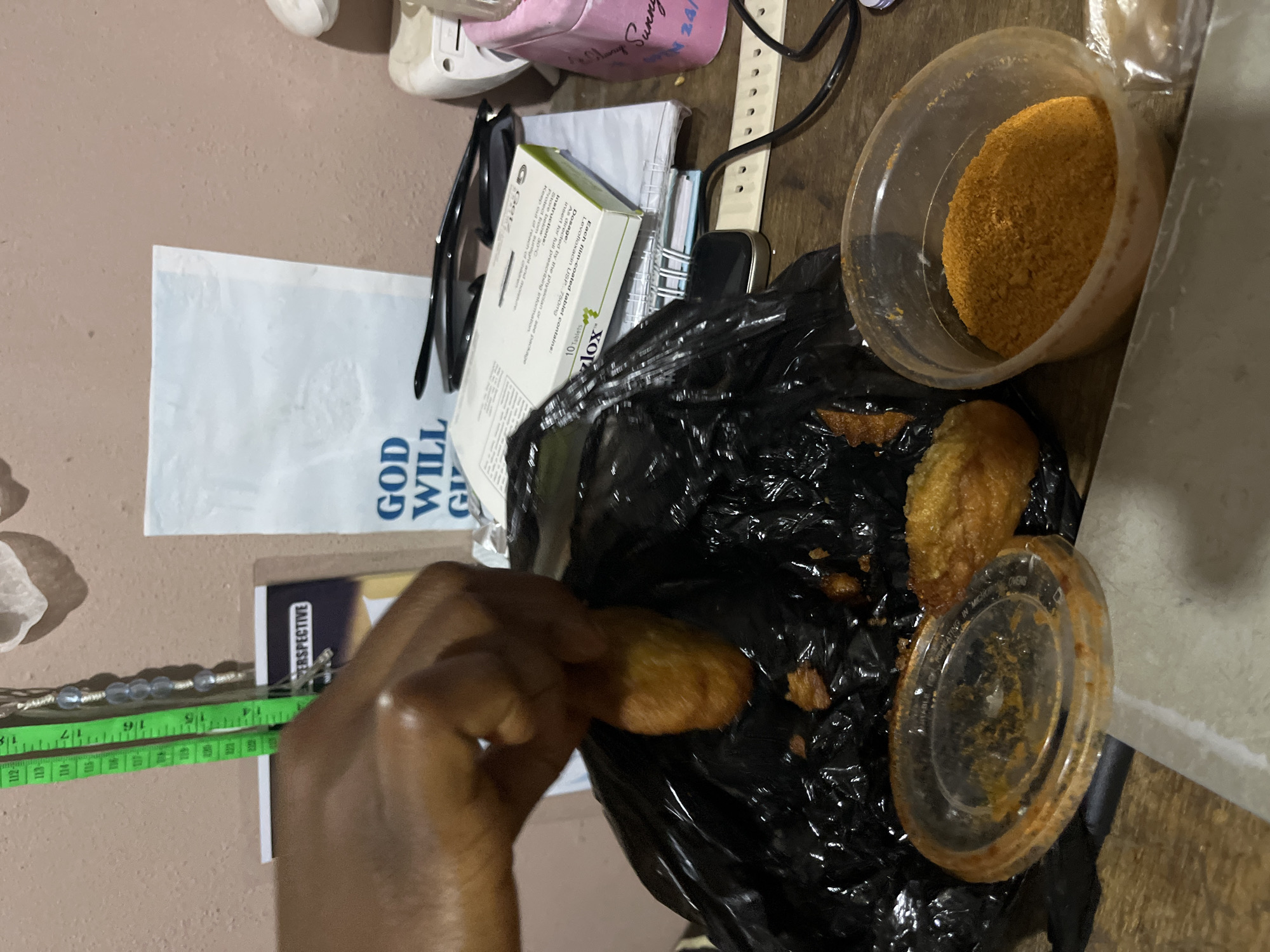The image depicts a close-up view of a table surface in what appears to be a home or personal space, likely in Rafin Par, Nigeria, given the provided context. In the foreground, a person's hand, with dark skin tone, is reaching into a black plastic bag, grasping a piece of fried food, possibly a plantain or yam. The bag contains several other pieces of similar fried food. To the left of the bag, there is a clear plastic container with a lid, stained with some orange residue, suggesting it previously held a sauce or seasoning. To the right of the bag, a clear plastic bowl is filled with a bright orange, granular substance, likely a spice or seasoning.
In the background, several objects are visible on the table. A white paper with blue text that reads "GOD WILL GIVE" is partially visible. Next to it, a pair of black-rimmed eyeglasses rests on top of a stack of papers, one of which is a box for medication labeled "Zyloc". A light-colored smartwatch with a dark strap is also present. Further in the background, a green measuring tape is hanging vertically, displaying measurements in centimeters, with numbers like 112, 113, 114, 15, and 16 visible. A wall with a peachy-pink hue serves as the backdrop. The overall scene suggests a casual, everyday setting, possibly during a meal or snack preparation.
