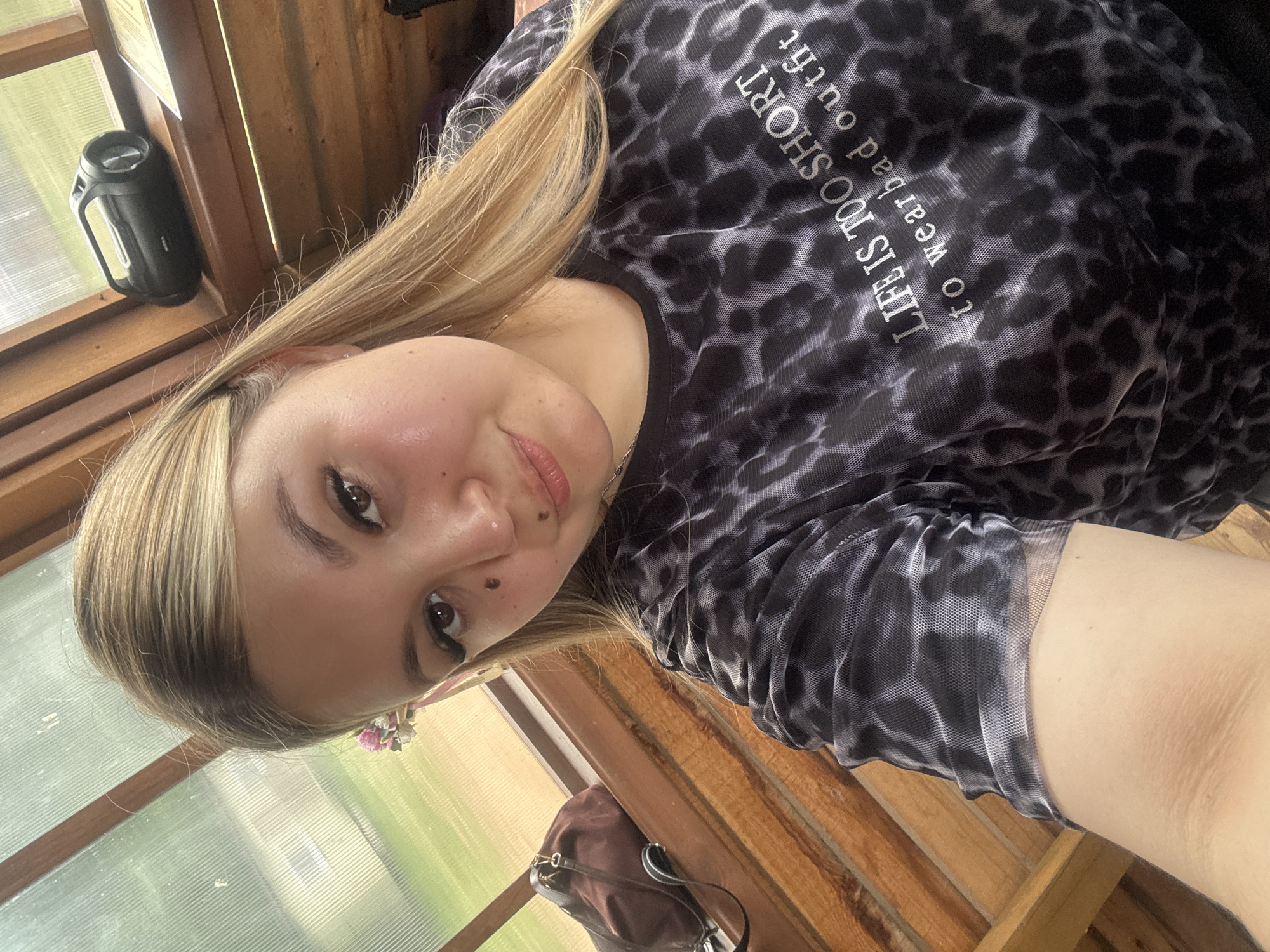 A young woman with blonde hair and makeup is taking a selfie. She is wearing a black and white leopard-print mesh shirt with the text "LIFE IS TOO SHORT to wear bad outfit" printed on it. A floral hair accessory is visible in her hair. Behind her, a brown bag rests on a wooden surface, and a black portable speaker sits on a shelf. The environment appears to be an enclosed wooden structure with a translucent roof, possibly a gazebo or a greenhouse, with greenery visible outside. The overall lighting suggests it is daytime.