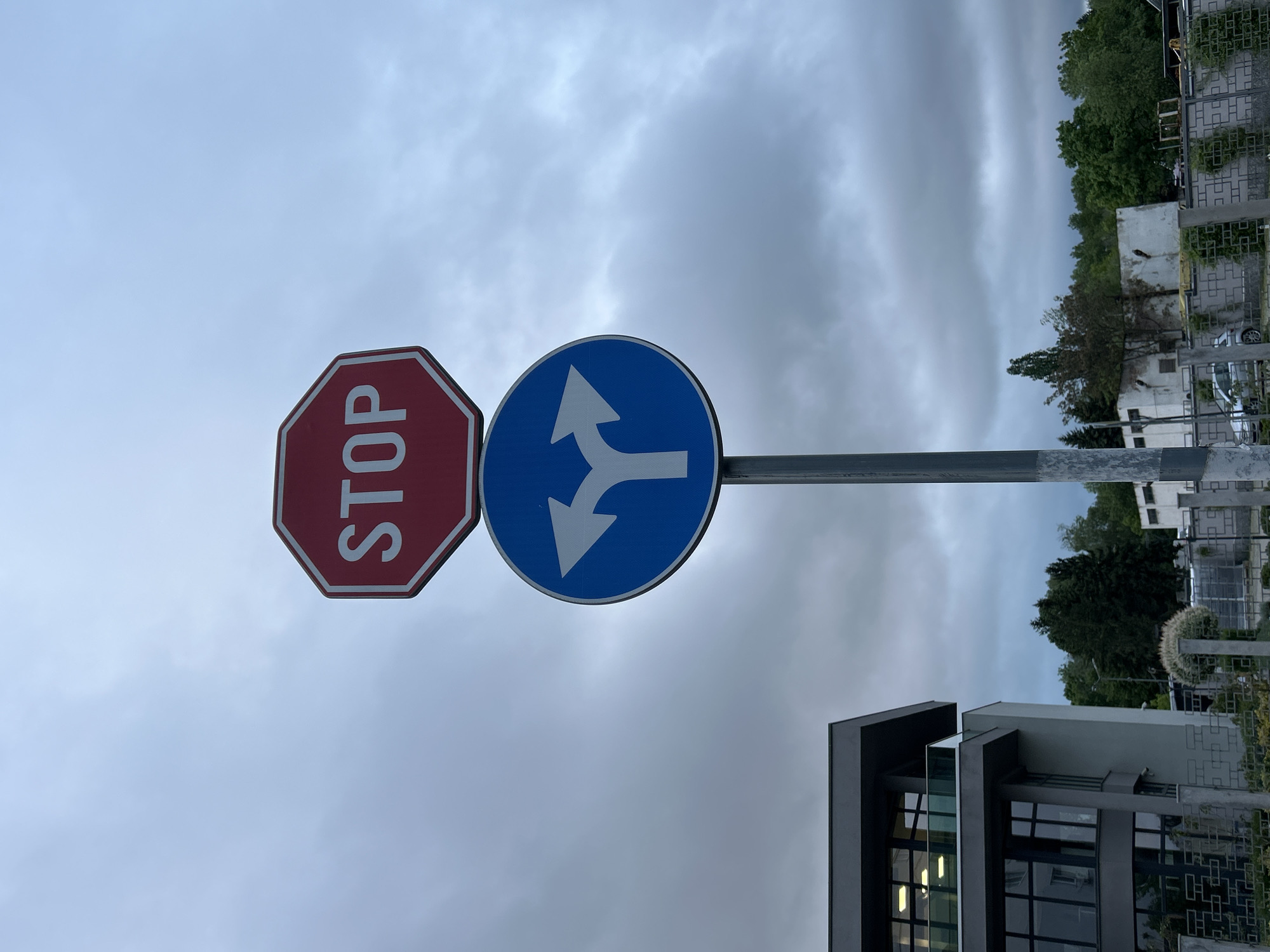 A stop sign and a blue roundabout direction sign are attached to a gray metal pole against a cloudy sky. In the background are trees, buildings, and a fence with square patterns. Part of a modern building with large windows is visible in the foreground. The location is the city of Unknown, Unknown.