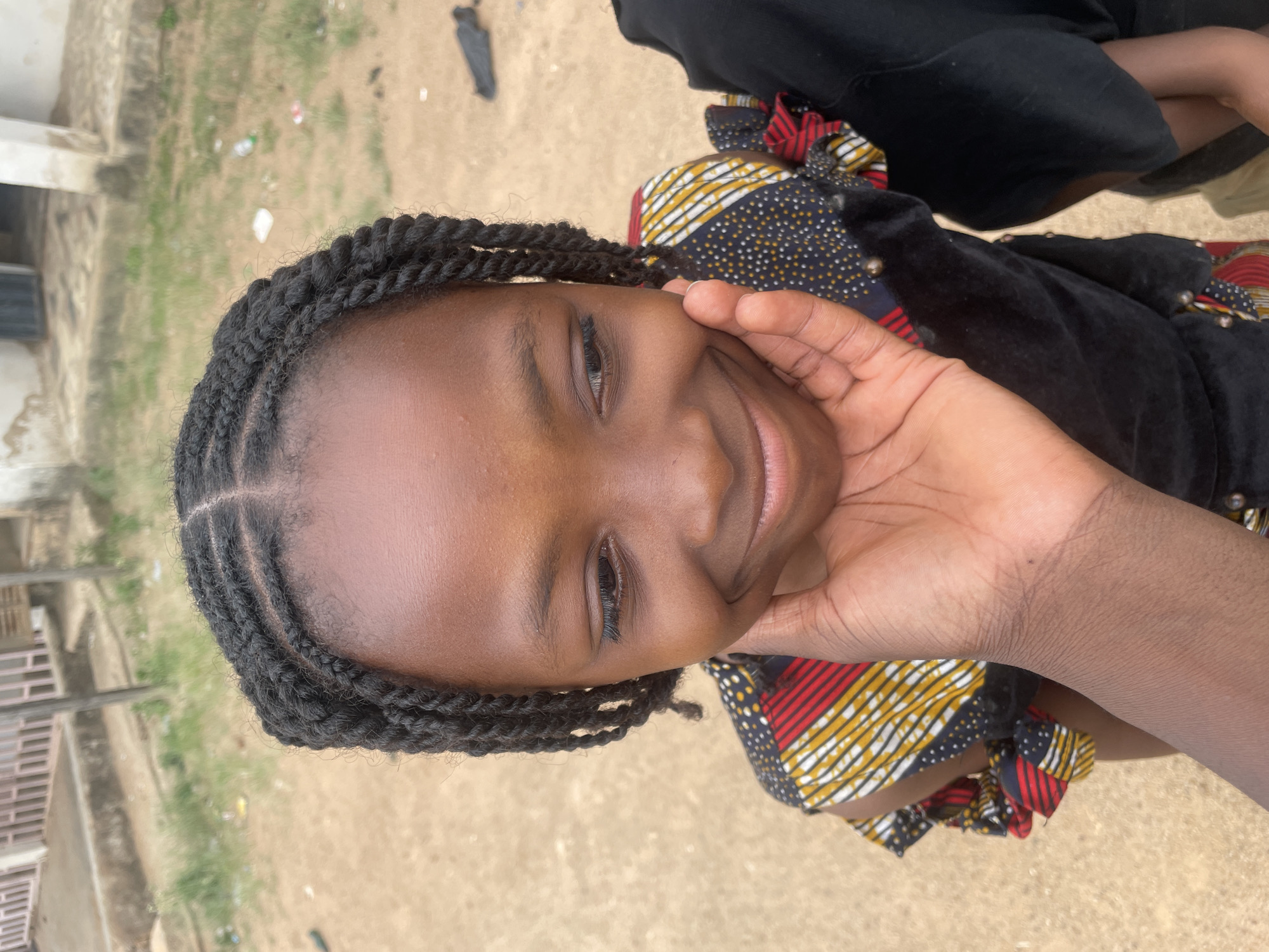 A close-up, vertical portrait captures a young person, likely a girl, with dark skin, smiling warmly and looking directly at the camera. Her head is tilted slightly, and her right hand rests gently on her cheek and chin. Her hair is styled in numerous neat, dark braided twists.
She is wearing a dark top or dress, accented with vibrant, patterned fabric visible on the sleeves or collar. This fabric features bold stripes of red, yellow, and white, interspersed with sections of white dots on a dark blue or black background.
The setting appears to be outdoors during the daytime, under natural light. The ground visible in the background is a light brown, earthy surface with sparse green vegetation and a few small pieces of litter. To the left, a partial view of a light-colored building with a dark railing is visible. The subject's expression is one of happiness and contentment. No text is visible in the image. The location context is Barakin Akawo, Nigeria.