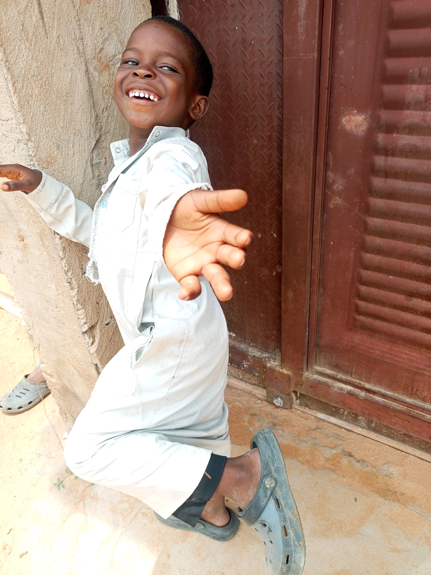A young boy with dark skin and short, dark hair kneels on a sandy surface next to a rough, textured wall. He is wearing a light-colored, loose-fitting garment that appears to be a traditional outfit, and dark-colored clogs on his feet. His face is turned towards the camera, and he is laughing with his mouth open, revealing white teeth. His right arm is extended forward, with his hand open and facing the camera, as if reaching out. To the right of the boy is a reddish-brown door with vertical ribbed panels. The ground is a mix of sand and some small patches of green vegetation. In the background, to the left of the boy, another light-colored shoe is visible on the ground. The lighting suggests it is daytime, with shadows indicating the sun is present. The overall scene depicts a candid moment of a joyful child.