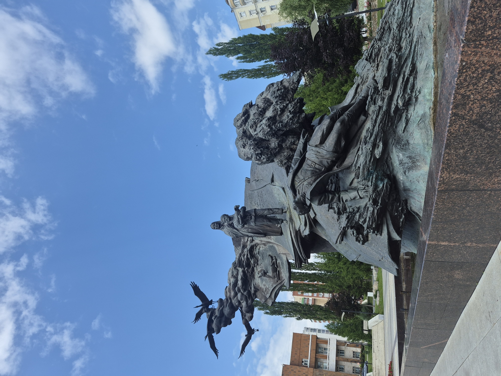 This is a full-shot, low-angle, exterior, eye-level, natural light, medium distance, daytime photograph of the Monument to the Friendship of Russia and Bashkiria in Ufa, Russia. The monument depicts a woman and child standing above a fallen soldier, with several bronze cranes flying above them. The monument is made of dark bronze and is set in a plaza with a fountain and a granite base. In the background, there are modern buildings, tall trees, and a blue sky with white clouds. The monument is a significant landmark in Ufa, commemorating the union of the Bashkir people with Russia.