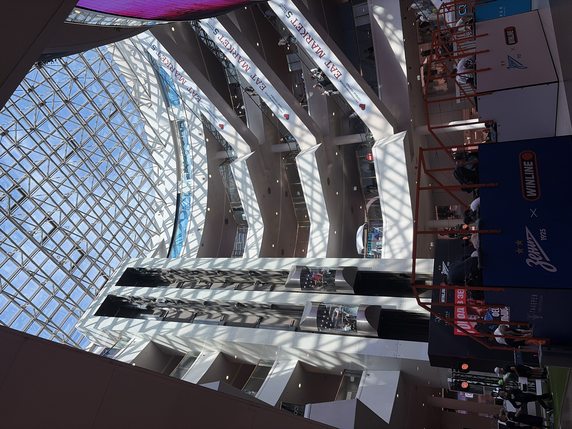 The image shows the interior of a multi-level shopping mall with a large glass atrium roof. The mall features escalators, elevators, and various retail levels. On the ground floor, there is a stage setup with branding for "Zenit" and "Winline", suggesting an event or promotion related to sports or gaming. Several people are visible on the escalators and on the stage area, some appear to be spectators or participants. The architectural style is modern, with clean lines and white surfaces. The lighting suggests it is daytime, with sunlight streaming through the glass roof. This scene is likely within the Galeria shopping mall in Saint Petersburg, Russia, which is known for its large atrium and modern design.