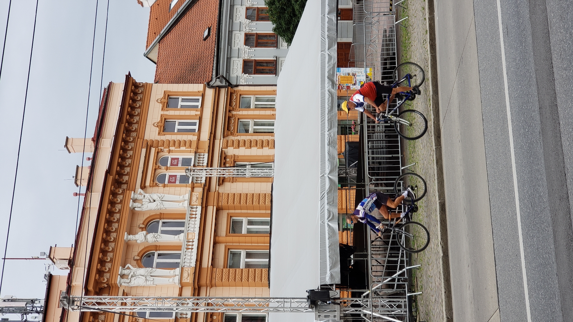 Two individuals are cycling on a paved walkway in front of a large, ornate orange building. The building features multiple stories with arched windows and decorative statues. A large white tent is erected in front of the building, extending across a significant portion of the frame. Metal barricades are positioned along the edge of the walkway. The cyclists are wearing athletic attire. The foreground shows a street with a white line marking. The scene appears to be outdoors in a city. This image was taken on Wednesday, July 19, 2023, on Hlavná Street in Prešov, Slovakia.