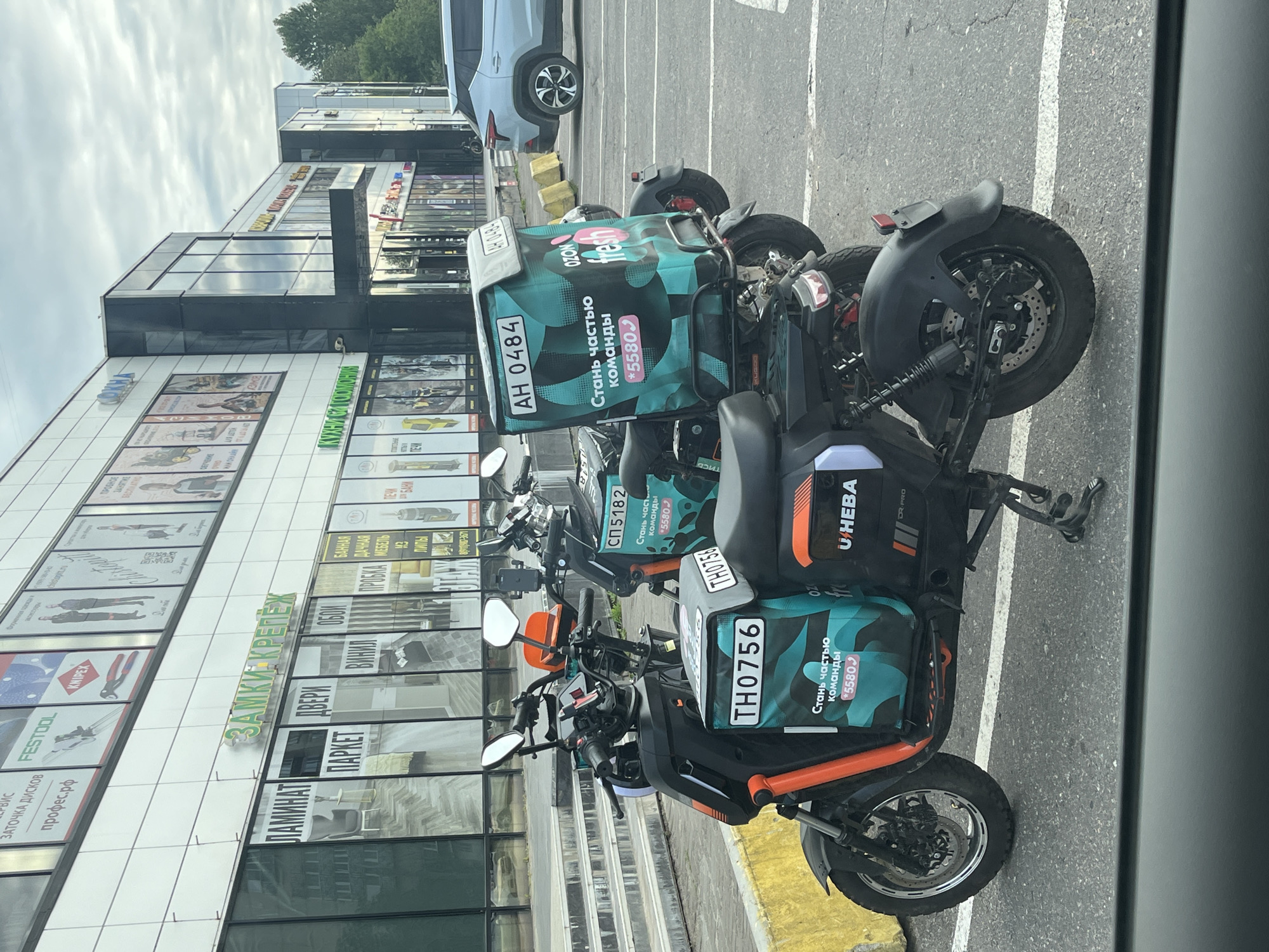 A group of Ozon delivery scooters are parked in front of a shopping center in Saint Petersburg, Russia. The scooters are black with orange accents and have large, teal-colored delivery boxes on the back. The boxes feature the Ozon logo and the text "Стань частью команды" (Become part of the team) and a phone number. The shopping center has large windows and signs with Russian text, including "ЗАМКИ КРЕПЕЖ" (Locks and Fasteners). A silver SUV is parked to the right of the scooters. The sky is overcast.