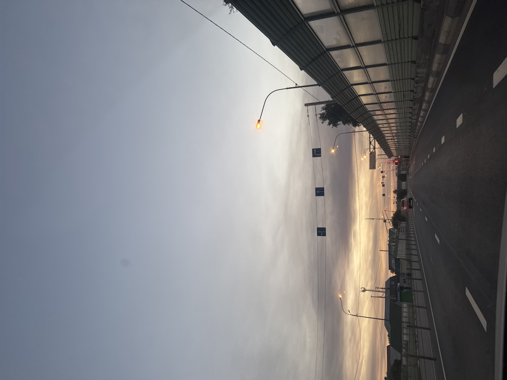 This image depicts a road in Mstikhino, Russia, at dusk or dawn. The sky is a pale blue with wispy clouds, transitioning to warmer hues of orange and yellow near the horizon. A road stretches into the distance, marked with white dashed lines. On the right side of the road is a tall, green sound barrier constructed of panels. Streetlights with orange lamps are visible along the roadside, with some illuminated. Traffic signs, including directional arrows, are suspended over the road on wires. On the left side, there are buildings and houses with green roofs, and a cellular tower is visible in the distance. A single vehicle, a silver SUV, is driving away from the camera on the right lane of the road. The overall atmosphere is serene, with the dimming light suggesting either the end of the day or the beginning of a new one.
