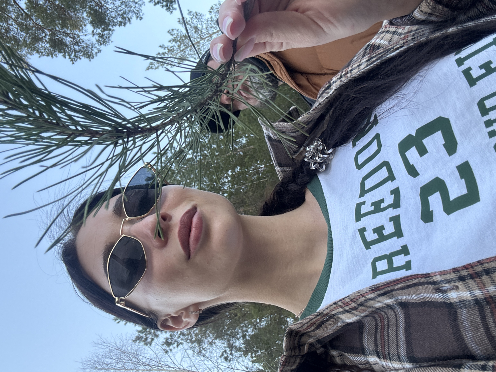 A low-angle shot shows a woman outdoors, holding a pine branch in front of her face. She wears sunglasses with gold frames. The woman has dark hair, partly braided and adorned with a silver hair accessory. She wears a white t-shirt with green lettering that says "REBEL 23". The shirt is layered under a plaid flannel shirt. She is also wearing a tan jacket. The background consists of trees and a blue sky. There is no evidence to identify the location in the city of Noviy, Russia.