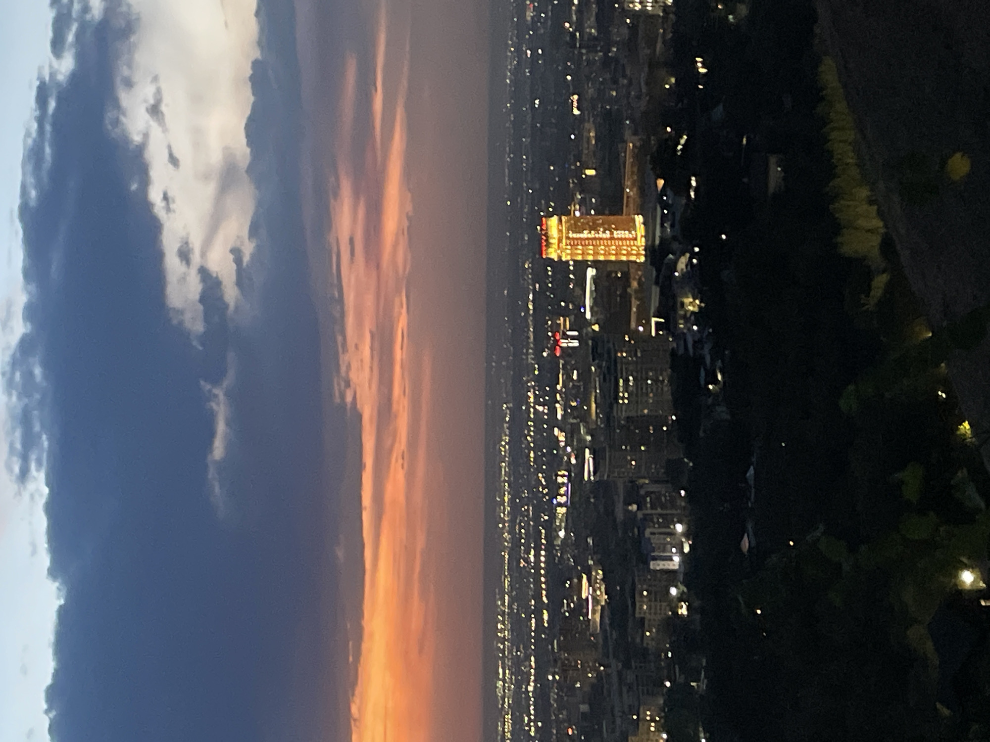 The image depicts a panoramic view of Almaty, Kazakhstan, at dusk. In the foreground, the dark silhouettes of trees and a slope with some lighting are visible. Beyond this, the city sprawls with numerous buildings and lights twinkling in the distance. A prominent, illuminated tall building with a golden glow stands out in the mid-ground, likely a landmark. The sky above is a dramatic canvas of twilight colors, with shades of dark blue, purple, and orange from the setting sun, interspersed with scattered clouds. The overall impression is a city transitioning from day to night, with the urban landscape illuminated against the darkening sky.