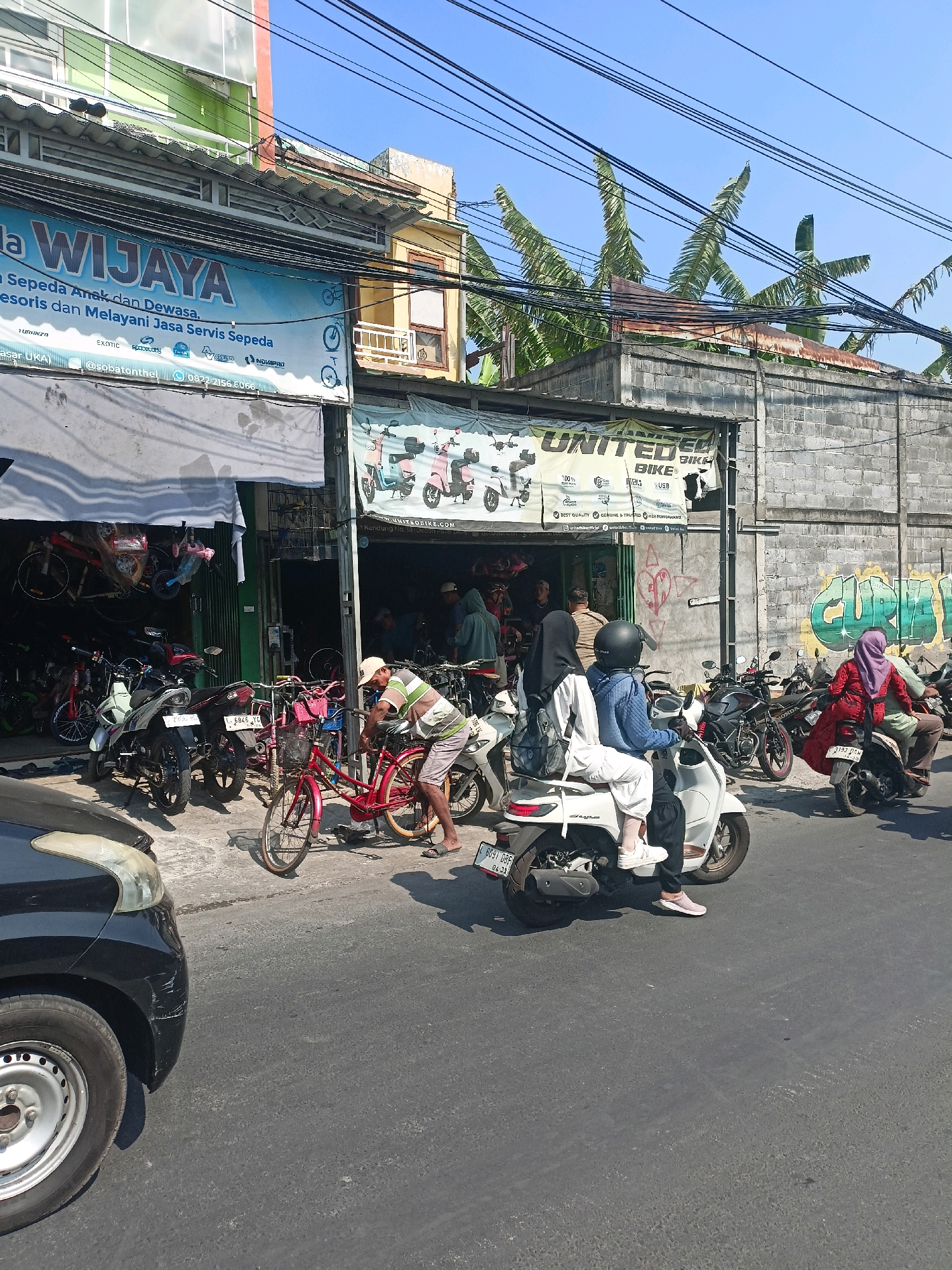 In Surabaya, Indonesia, a street-level view captures a bustling scene outside a bicycle shop named "WIJAYA" and "UNITED BIKE". A blue banner with "WIJAYA" in large white letters is prominently displayed on the building's facade, advertising "Sepeda Anak dan Dewasa. Aksesoris dan Melayani Jasa Servis Sepeda." (Bicycles for Children and Adults. Accessories and Serving Bicycle Repair Services). Below this, there are various logos, including "TURANZA", "EXOTIC", "PACIFIC", and "UNITED".
The storefront itself features an open entrance with several bicycles hanging inside, suggesting a retail space. Outside the shop, motorcycles and bicycles are parked along the roadside, indicative of a busy commercial area. A man in a striped shirt and shorts is working on a red bicycle.
Several people are present, with some on motorcycles. Two individuals are seated on a white scooter in the foreground, one driving and the other a passenger. Another motorcycle, black in color, is parked nearby, with a person wearing a red hijab seated on it. A black car is parked on the left side of the image, its front bumper showing some scratches.
The environment appears to be a typical urban street with a clear blue sky overhead, interspersed with numerous electrical wires. The buildings have a mix of architectural styles, with some featuring brick walls and others more modern facades. Banana trees are visible behind the buildings, adding a touch of tropical vegetation to the urban landscape. Graffiti is present on the brick wall to the right of the "UNITED BIKE" shop. The road surface is asphalt and appears somewhat worn.