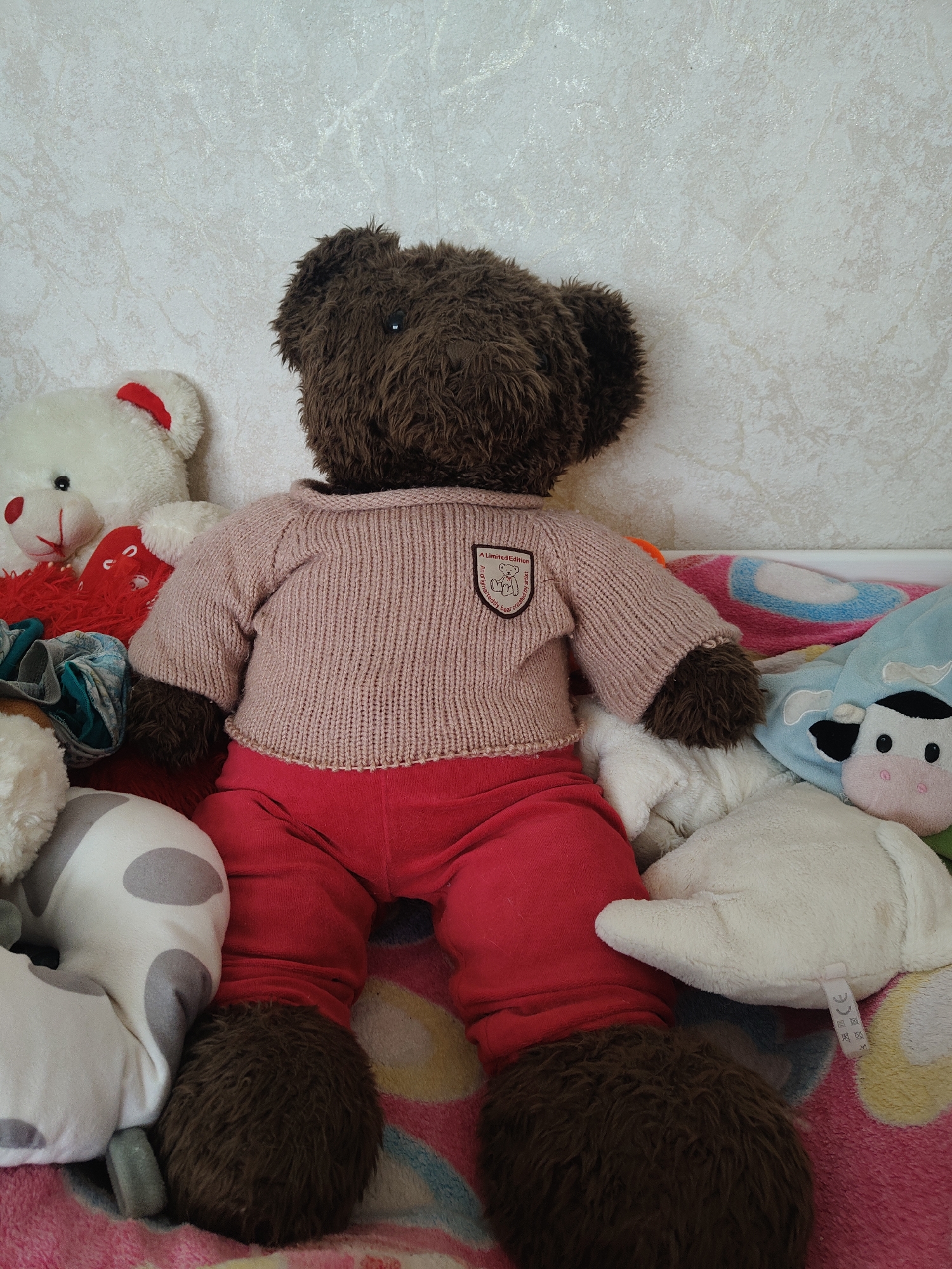 A dark brown teddy bear is sitting upright, facing away from the camera, wearing a light pink knitted sweater and red pants. The bear is surrounded by other stuffed animals, including a white teddy bear to its left with red accents, a white stuffed pillow with grey spots, and a light blue plush toy with a cow face. The stuffed animals are arranged on a pink blanket with a colorful pattern. The background is a textured wall with a light, neutral color. The scene appears to be indoors, in a domestic setting. There is no indication of the specific location within Kovalevskoye, Russia, or any actions or interactions beyond the arrangement of the objects.