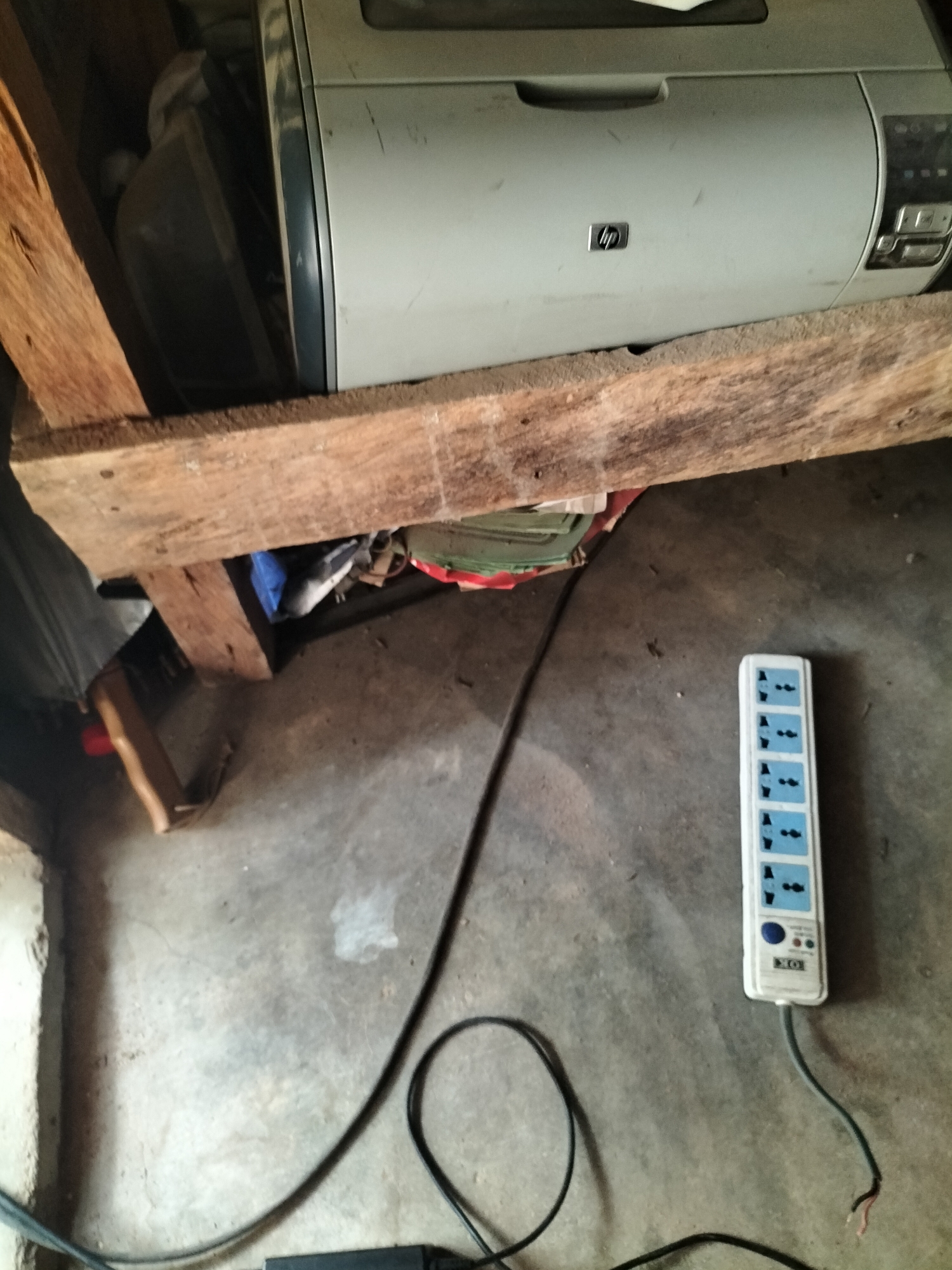 An overhead shot captures a cluttered storage area, possibly in Jalingo, Nigeria. A grey HP printer rests precariously on rough-hewn wooden planks, its surface dusty. Below the printer, a jumble of items, including a blue bag and green fabric, are visible. On the concrete floor, a white multi-socket power strip with six blue outlets and two control buttons is plugged into an electrical cord. Another black electrical cord snakes across the floor, its end frayed, suggesting disrepair. The environment appears unkempt, with dust and debris present on the floor and surfaces. The lighting is dim, casting shadows and contributing to the overall impression of a neglected space.