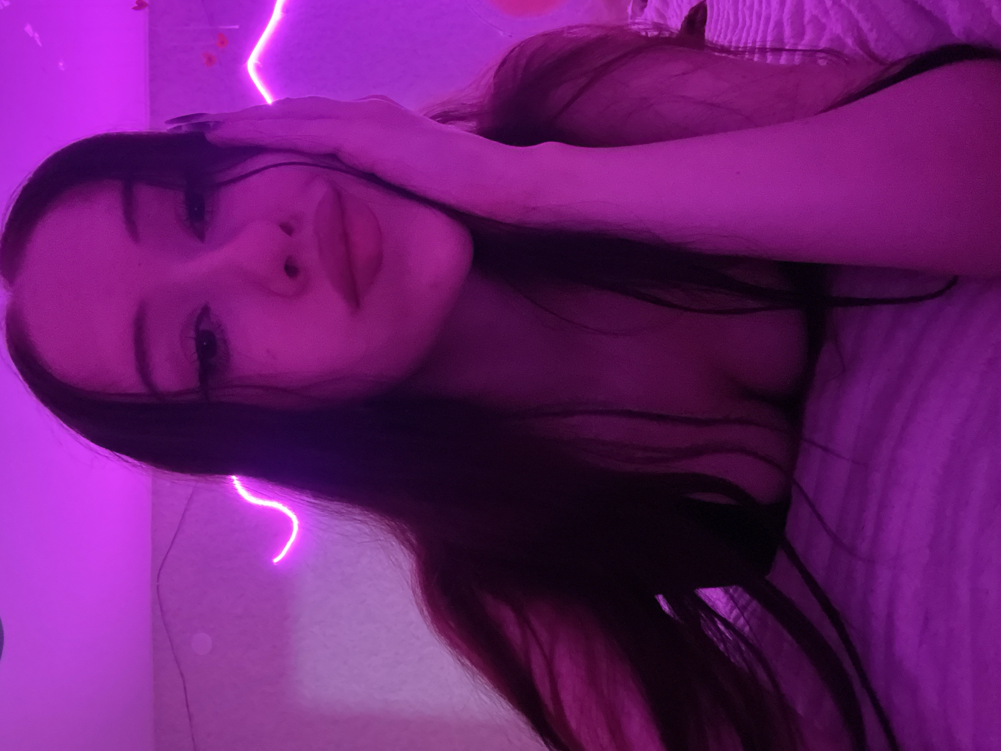 This is a medium shot, close-up selfie of a young woman lying down, likely in a bedroom. The lighting is predominantly pink and purple neon, creating a moody and atmospheric effect. The woman has long dark hair, fair skin, and is wearing dark clothing that reveals her décolletage. She is looking directly at the camera with her hand resting on her cheek, and her lips are parted. The background shows a wall with pink neon lights, suggesting a decorative element. The woman is positioned on a bed with a textured surface. There are no discernible elements in the image that would indicate a specific location in the city of Votkinsk, Russia.