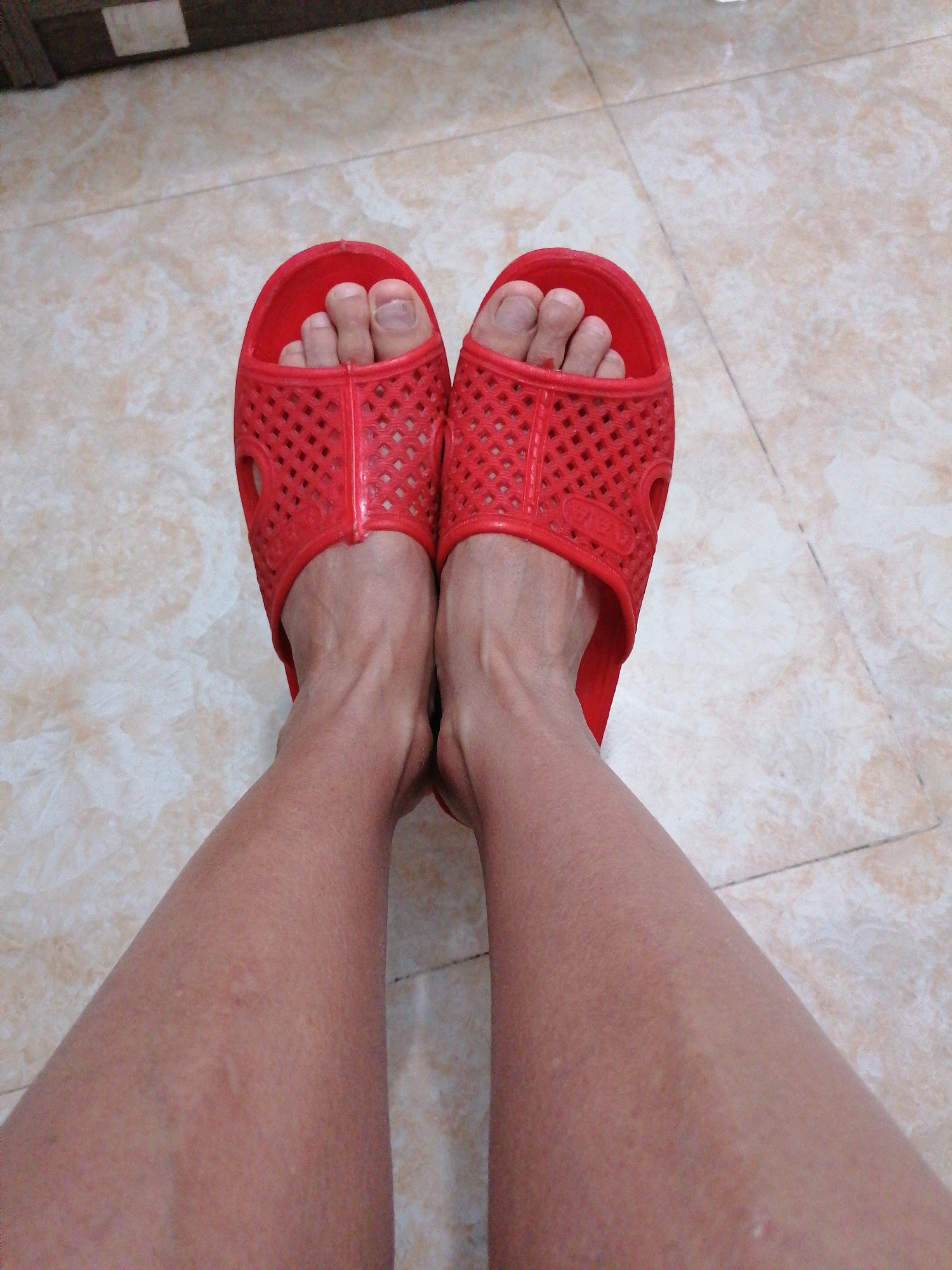 The image displays a first-person view of a person's lower legs and feet, adorned with a pair of bright red plastic slide sandals. The setting appears to be indoors, with the person standing or sitting on a light-colored tiled floor featuring a subtle, variegated pattern.
The sandals are a prominent feature, made of a vibrant red plastic material with a distinctive perforated design, characterized by small diamond-shaped cutouts covering the upper strap and sides. The person's toes are visible through the front opening of the sandals, and their toenails are natural and unpolished. The skin on the feet and lower legs shows natural texture, with some visible veins, particularly around the ankles. Several small bumps or marks are discernible on the shins and calves, suggesting skin imperfections or possibly insect bites.
A small, faded white mark, likely an illegible brand name or logo, is visible on the inner side of the right sandal (on the viewer's left foot). No specific activity is taking place beyond the person simply wearing the footwear. The time of day and weather conditions are not discernible from the image. The tiled floor and casual footwear suggest a relaxed, domestic environment. This scene is set in Bai Chay Ward, Vietnam.